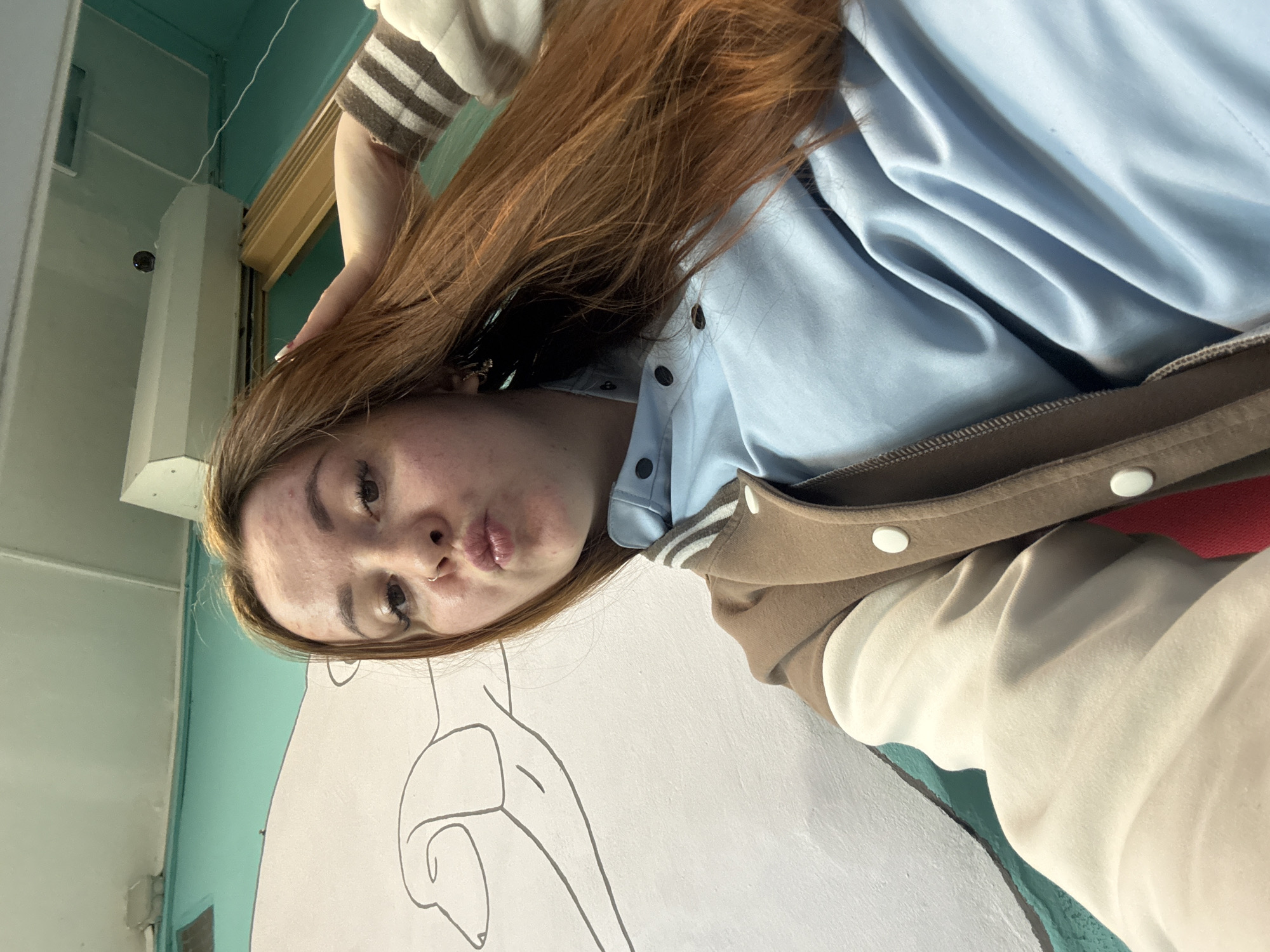 This media file is an angled selfie of a young woman taken indoors in Izhevsk, Russia. The woman has long, reddish-brown hair and is seen making a playful "duck face" expression with puckered lips, looking directly at the camera. She has a nose ring. She is wearing a light blue button-up shirt underneath a two-toned bomber jacket, which features a tan or brown body and lighter, possibly beige, sleeves. One arm shows a striped cuff.
The background reveals an indoor setting with walls painted in different colors: a prominent light blue-green or teal section and a white section. On the white wall, there is a large, stylized black line drawing that appears to be an abstract human form or anatomical outline. Architectural details like doorframes or columns are visible, and a small, dark, spherical object, possibly a security camera or light fixture, is mounted on a column in the upper left of the frame. The lighting is bright and even, suggesting it is daytime. There is no visible text in the image.