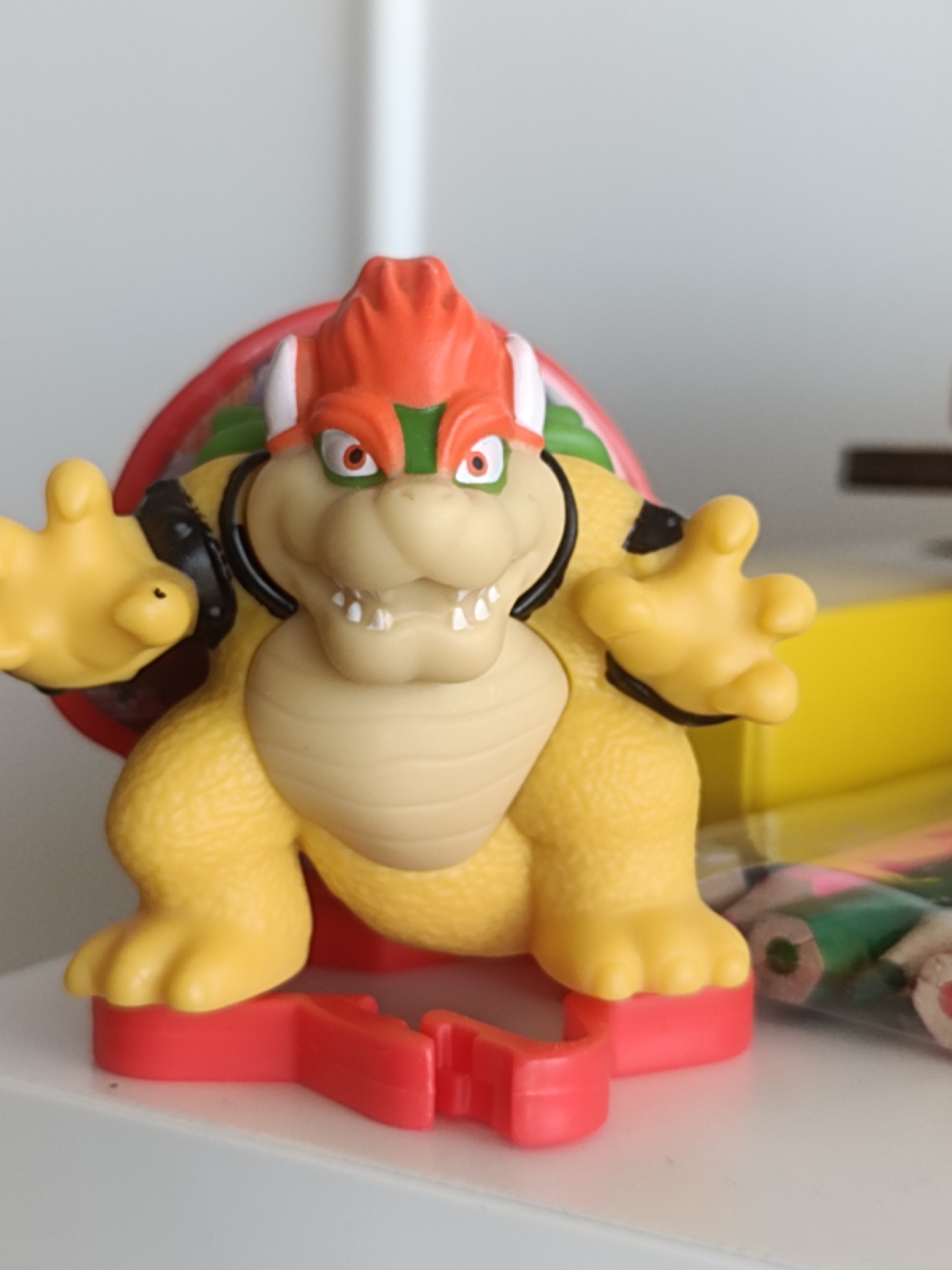 A bright yellow Bowser toy figure stands on a red plastic base. The toy has orange spikes on its head, green eyebrows, and red eyes with black pupils. It has large, yellow paws with three fingers and a thumb, and its body is covered in scaly yellow skin. The toy is positioned on a white surface, possibly a shelf, with a blurred background that suggests an indoor environment. To the right of the toy, a clear plastic bag filled with colored pencils is visible, also resting on the white surface. The lighting suggests a well-lit room, possibly with natural light coming from an unseen source. There is no visible indication of the city of Dubai, United Arab Emirates, in this image.