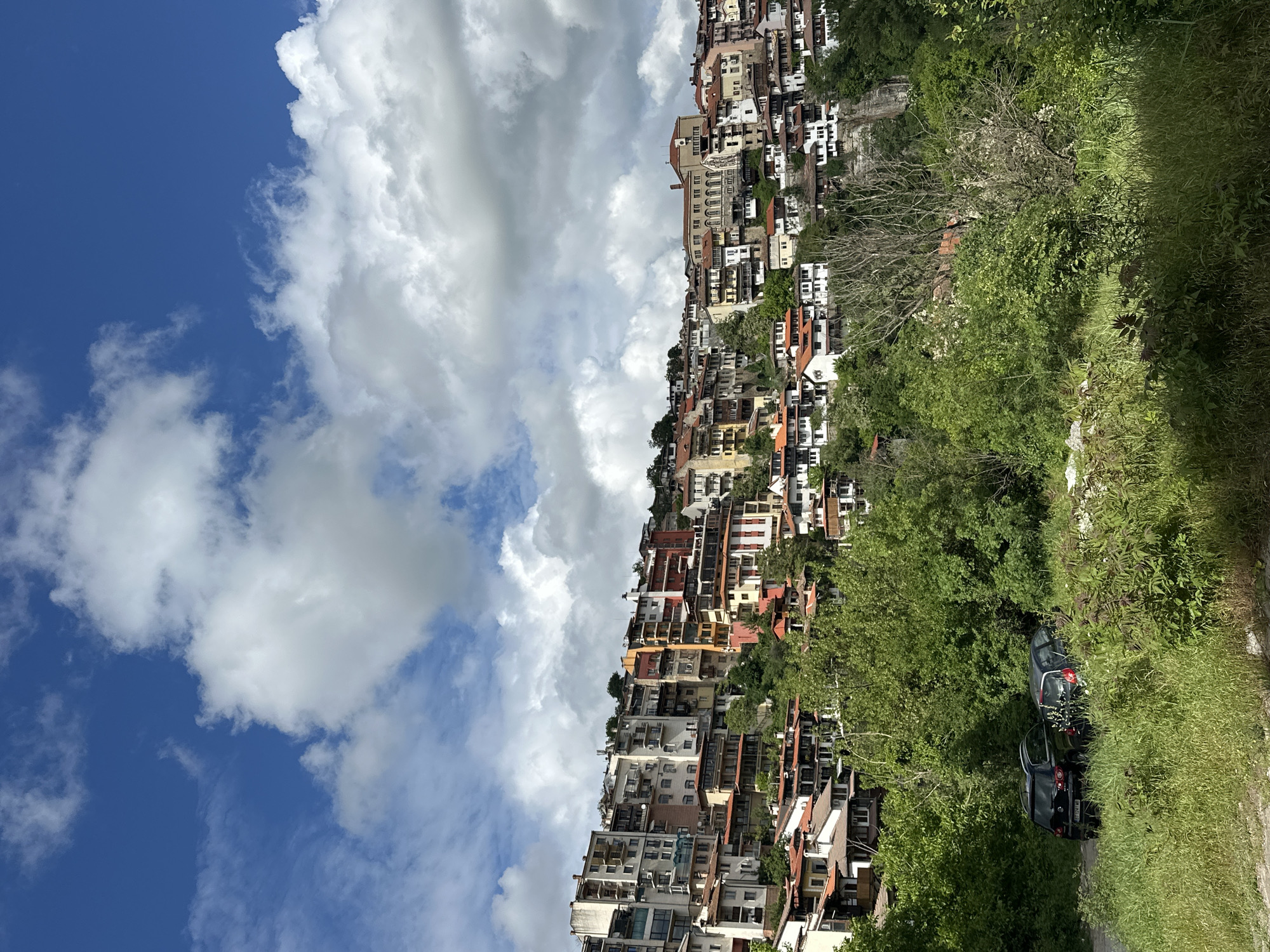 The image depicts a hillside residential area in Veliko Tarnovo, Bulgaria, under a partly cloudy sky. Numerous multi-story buildings of varying colors and architectural styles are clustered together, seemingly built into the slope of the hill. Green vegetation is prevalent, including trees and grass, bordering what appears to be a road. Two dark-colored vehicles are parked alongside the road. The sky is blue with white, puffy clouds.