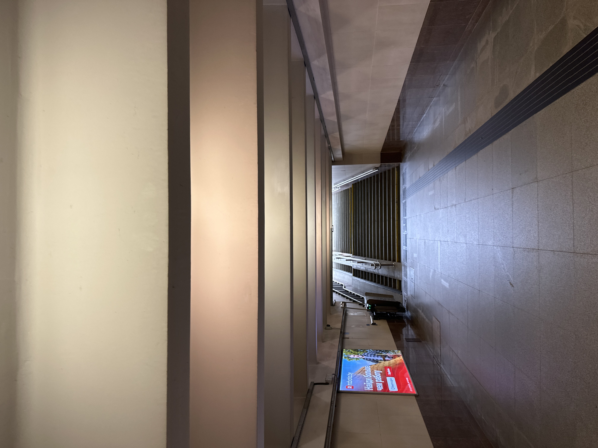 The image shows an underground pedestrian passage. It is illuminated by artificial lighting, creating a warm, soft glow. The passage is symmetrical, with a wide, tiled floor and beige walls. A series of parallel beams run across the ceiling, adding to the sense of depth. On the left side of the passage, a large illuminated advertisement for "robota.by" is displayed, featuring a man in a checkered shirt. Further down the passage, a set of stairs leads upwards, flanked by railings and a modern ticket barrier system. A dark tactile paving strip runs down the center of the floor, guiding pedestrians. The overall impression is one of a clean, well-maintained, and functional public space. This pedestrian underpass is located in Minsk, Belarus.