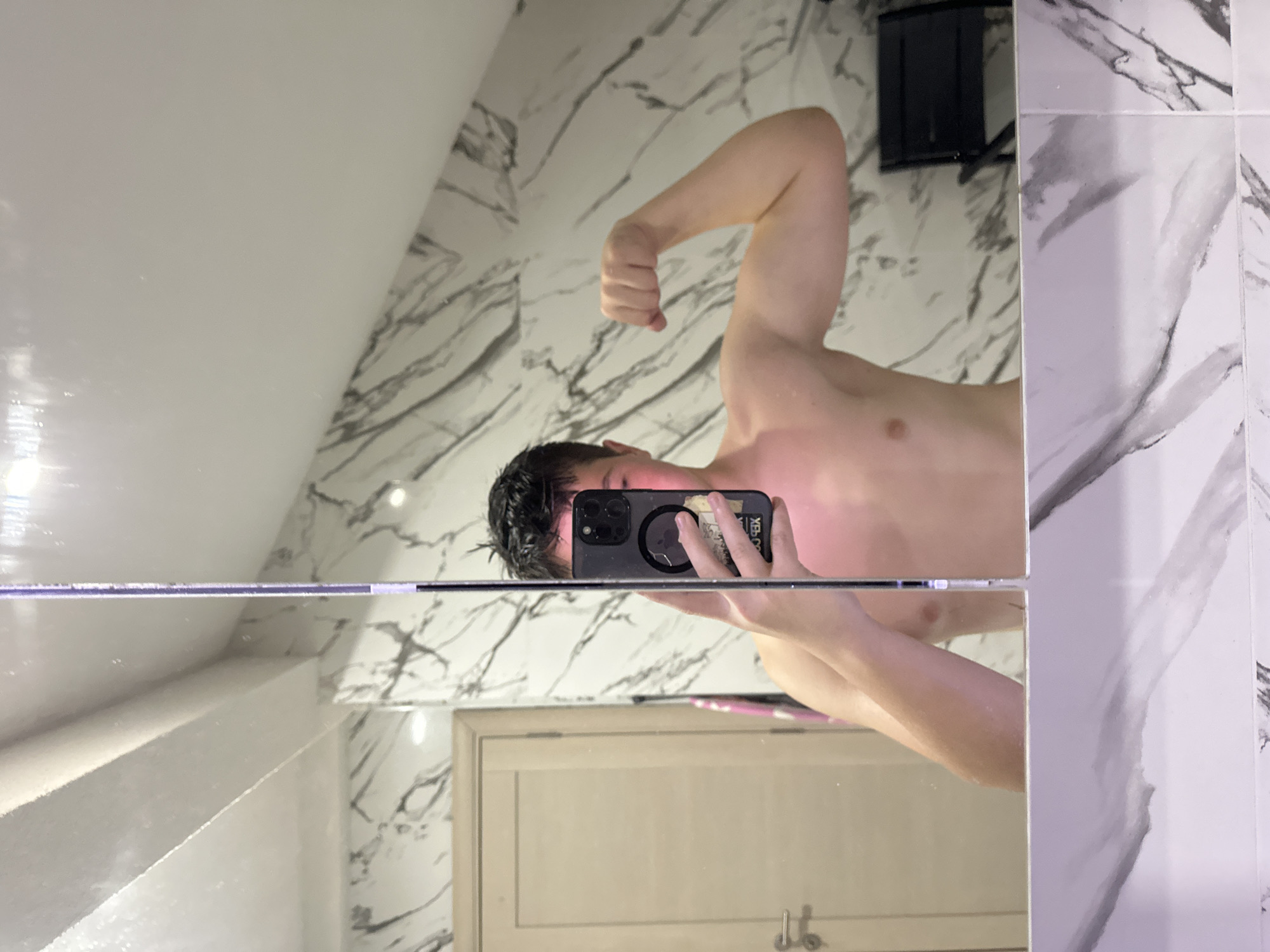 A shirtless man is taking a selfie in a bathroom mirror. He is flexing his right arm. The bathroom has white marble-patterned walls. There is a cabinet door visible in the mirror reflection. Based on the location, the bathroom could be within any residential or hotel property in Budva, Montenegro.