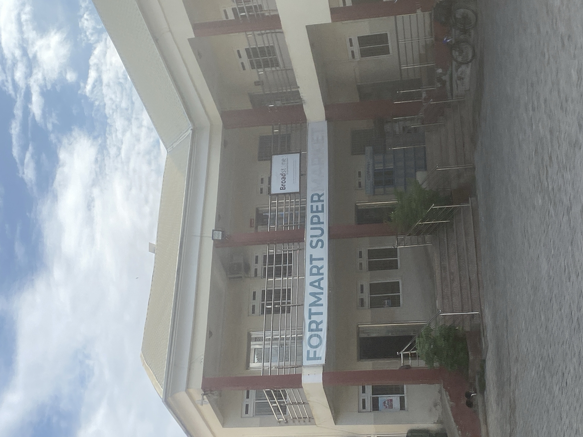 The image shows the exterior of the "Fortmart Super" supermarket in Angwan Sayawa, Nigeria. The building is a multi-story structure with a light beige facade, dark brown trim, and multiple windows. A large banner with the supermarket's name is vertically oriented on the building's front. A secondary sign for "Broadstone" is visible above the main sign. Metal railings and stairs lead to the building's entrance. A bicycle is parked near the entrance on the right. The sky is visible, with clouds partially obscuring it. The surrounding area is paved.