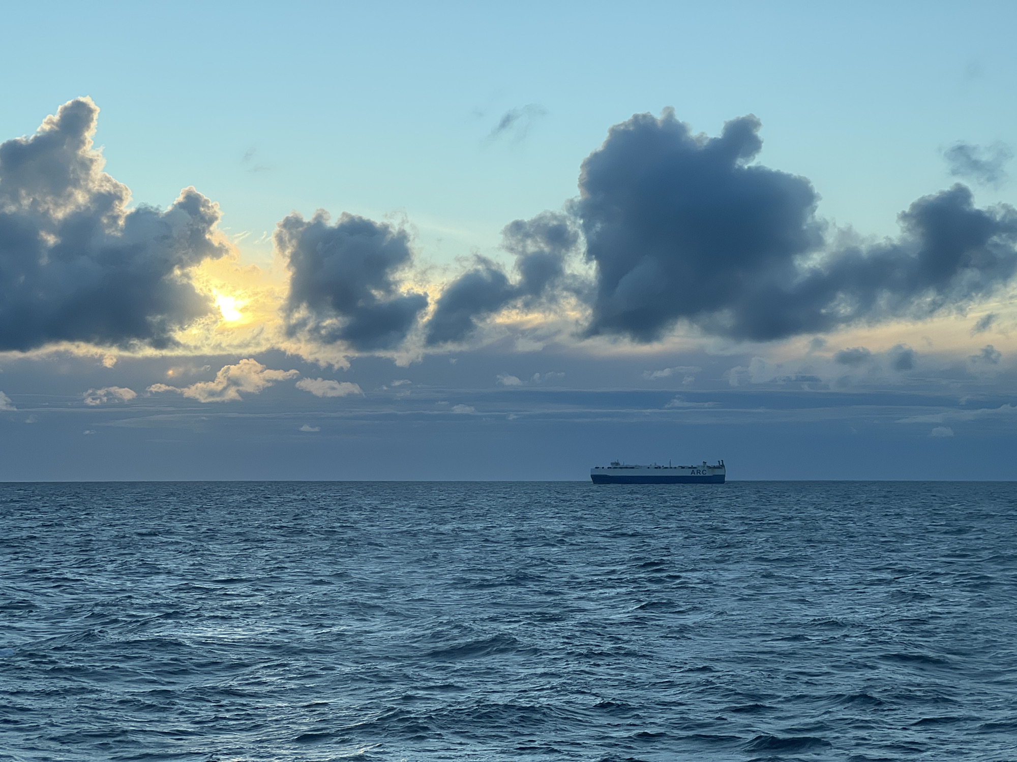 A cargo ship is visible on the horizon, sailing across a vast expanse of deep blue water under a dramatic sky. The sun is peeking through dark, textured clouds, casting a warm, yellow glow that contrasts with the cooler tones of the sky. The sea is slightly choppy, with small waves creating ripples across its surface. The overall scene suggests a serene, yet powerful natural environment. The location is unknown.