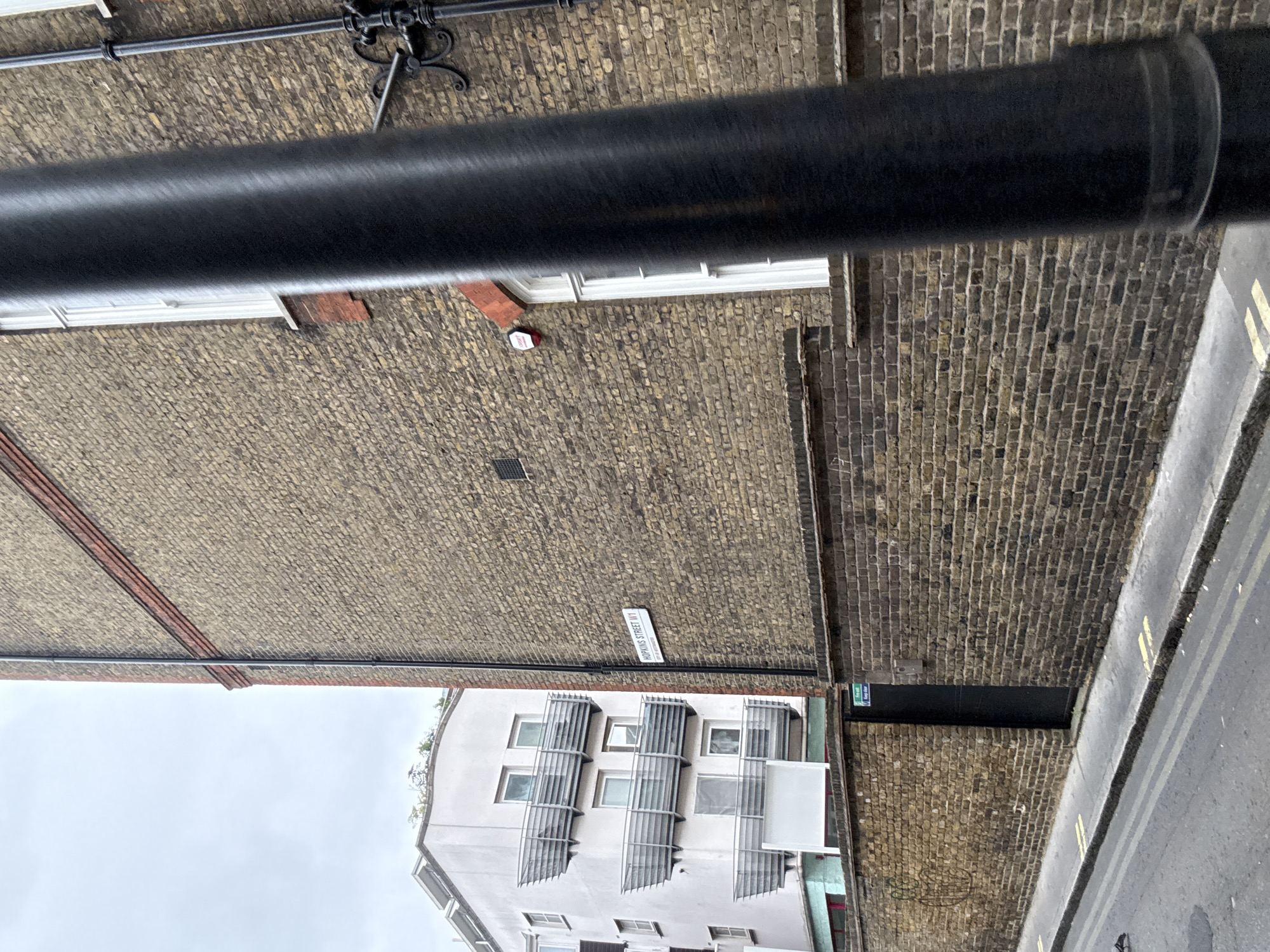 The image shows a street in London, United Kingdom. In the bottom-center of the frame is a modern, multi-story building with numerous windows and slatted sunshades. Above this building, a brick wall forms the backdrop. A street sign is visible on the brick wall, bearing the name "Hopkins Street W1". A black metal pipe is visible in the upper-middle ground. A section of road with painted yellow lines is visible on the right side of the image. The sky appears overcast.