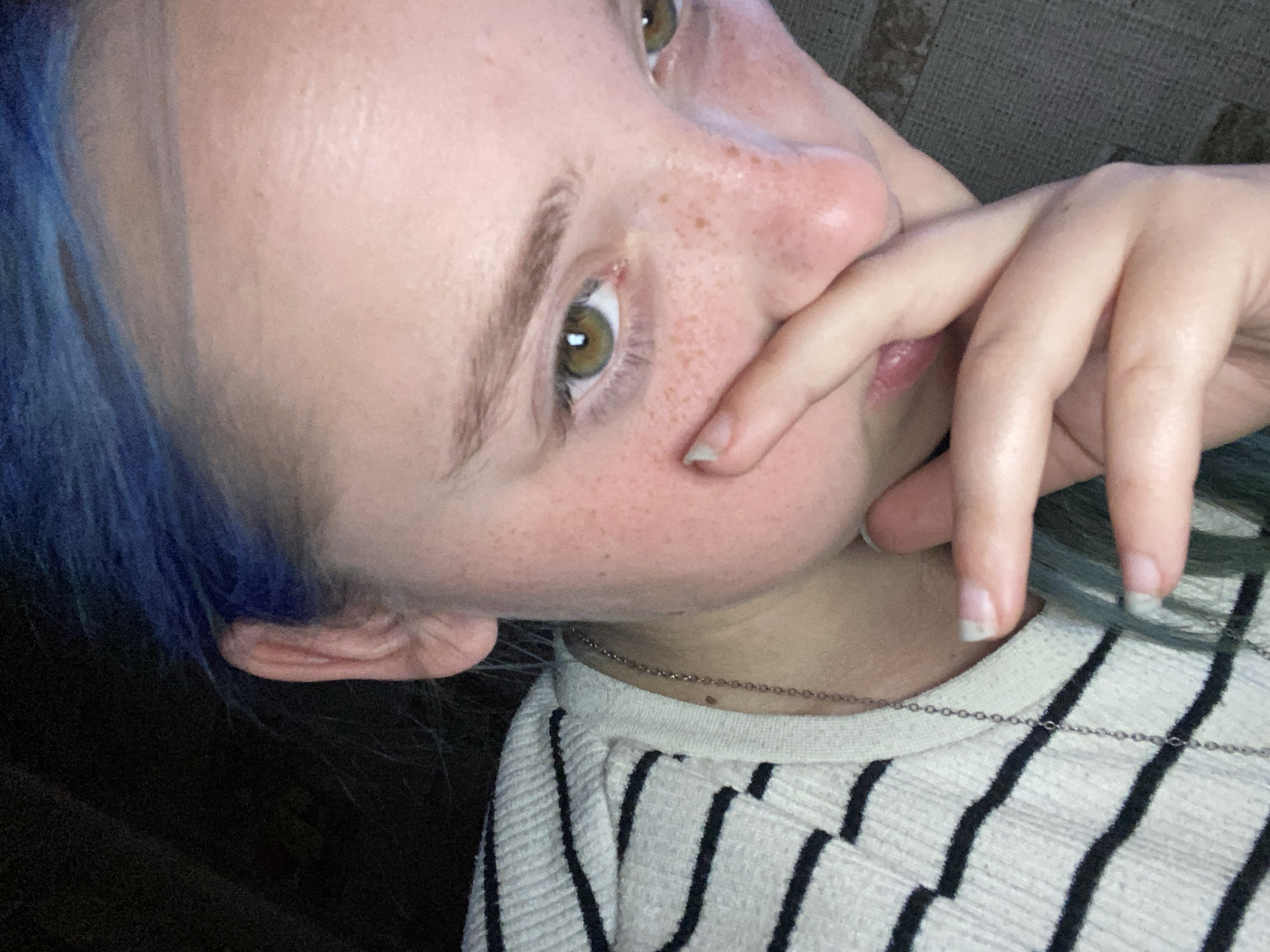 This is a close-up, eye-level, indoor, undated, indoor photograph of a person with blue hair. The person is facing slightly to the right, with their left hand covering their nose and mouth area. Their eyes are open and looking slightly to the left. The person is wearing a black and white striped sweater and a silver chain necklace. The background is dark and out of focus, with some texture that suggests a wall or fabric. The lighting is dim, creating shadows on the person's face. The image quality is moderate, with some noise and blur. The scene is not identifiable as being located in Nova Praha, Ukraine.