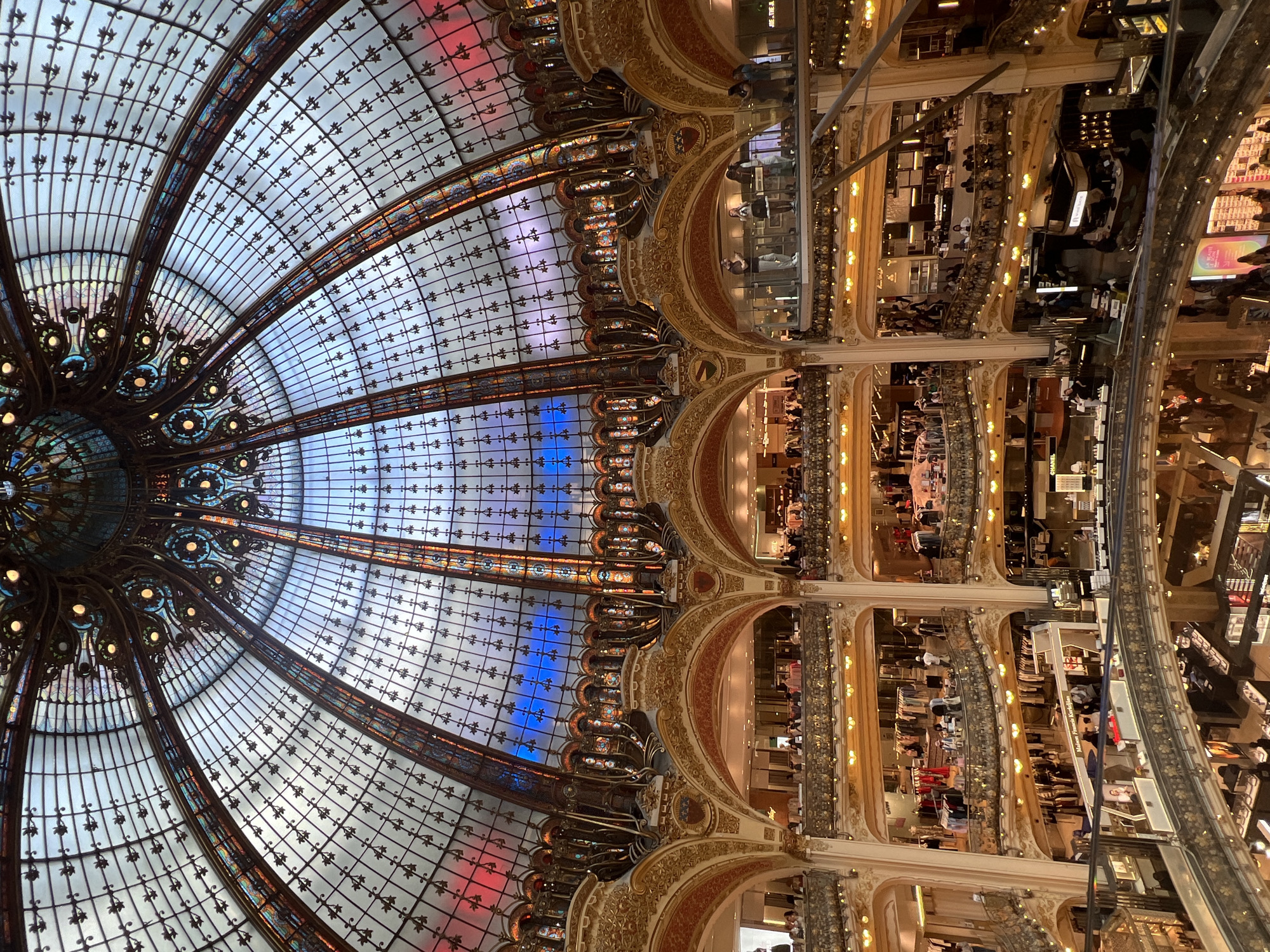 The image captures the ornate interior of the Galeries Lafayette Haussmann department store in Paris, France. The central focus is the magnificent Art Nouveau glass dome, a masterpiece of stained glass and ironwork, casting a soft, diffused light into the multi-story retail space below. Sunlight streams through the dome, illuminating its intricate patterns and colorful panels that transition from red to blue hues.
The department store features multiple levels, each visible with balconies adorned with decorative golden railings and lamps. Racks of clothing are displayed on various floors, with mannequins and shop displays visible. People are seen browsing the merchandise, walking along the balconies, and congregating in different areas of the store. Escalators and staircases connect the levels, facilitating movement within the bustling retail environment. The architecture is characterized by grand arches, elaborate moldings, and a sense of opulent grandeur. The overall scene depicts a vibrant, upscale shopping atmosphere within a historically significant architectural landmark.