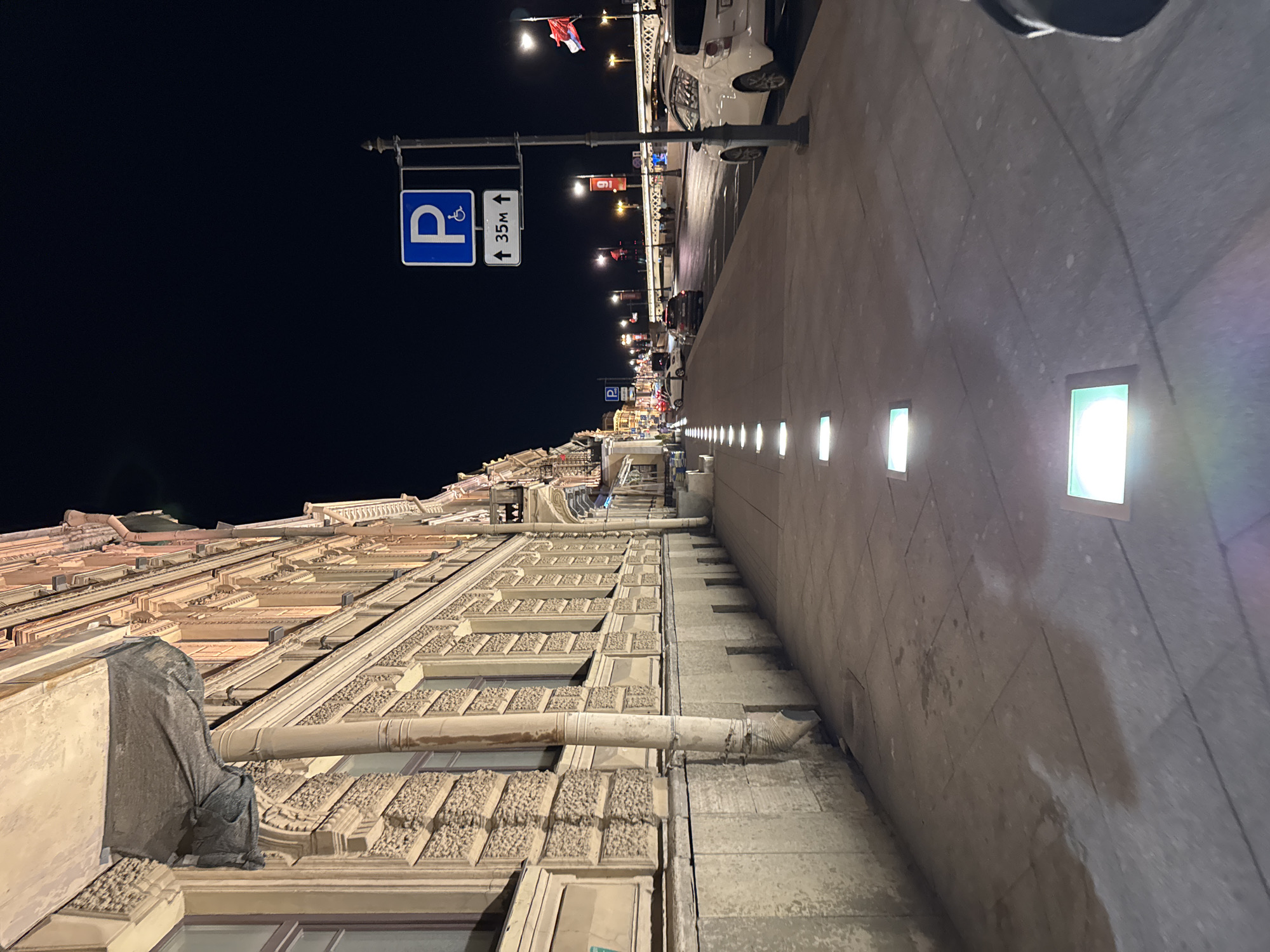 The image depicts a street scene at night in Saint Petersburg, Russia. The view is oriented vertically, with the building façade on the left, the sidewalk and street on the right, and a black sky above.
A tall, ornamented building with multiple windows and architectural details runs along the left side of the frame. A drainpipe is visible on the façade.
The sidewalk is paved with rectangular stones and features several inset square lights that illuminate the area. A street runs alongside the sidewalk, with parked cars visible.
A street sign with a wheelchair symbol and an arrow pointing upwards with "35M" written on it is mounted on a metal pole. Additional streetlights with flags are also seen. The street appears to stretch into the distance with illuminated buildings and streetlights.