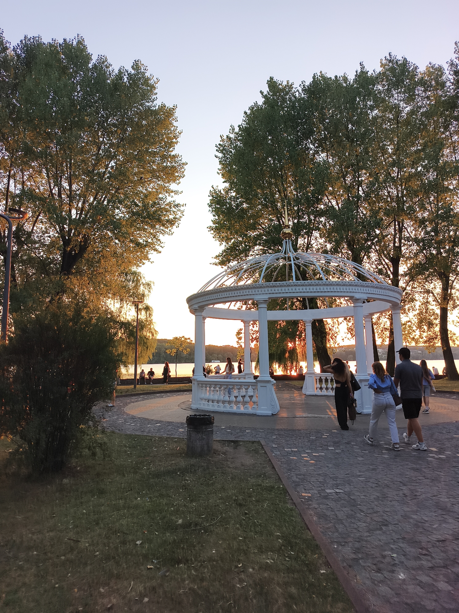 The image depicts an outdoor scene in Ternopil, Ukraine, likely during sunset, given the warm lighting. In the foreground, a paved walkway made of dark cobblestones curves towards a white, ornate gazebo with a domed roof and decorative balustrades. Several people are walking on the path near the gazebo; two women and two men are visible in the mid-right of the frame. Further back, a group of people are seated or standing along a waterfront, with trees lining the distant shore. Two tall lampposts are visible on the left side of the image, one partially obscured by a dense green bush. The sky is a pale orange, transitioning to clear blue, indicating the sun is low on the horizon. The overall atmosphere suggests a park or promenade area.