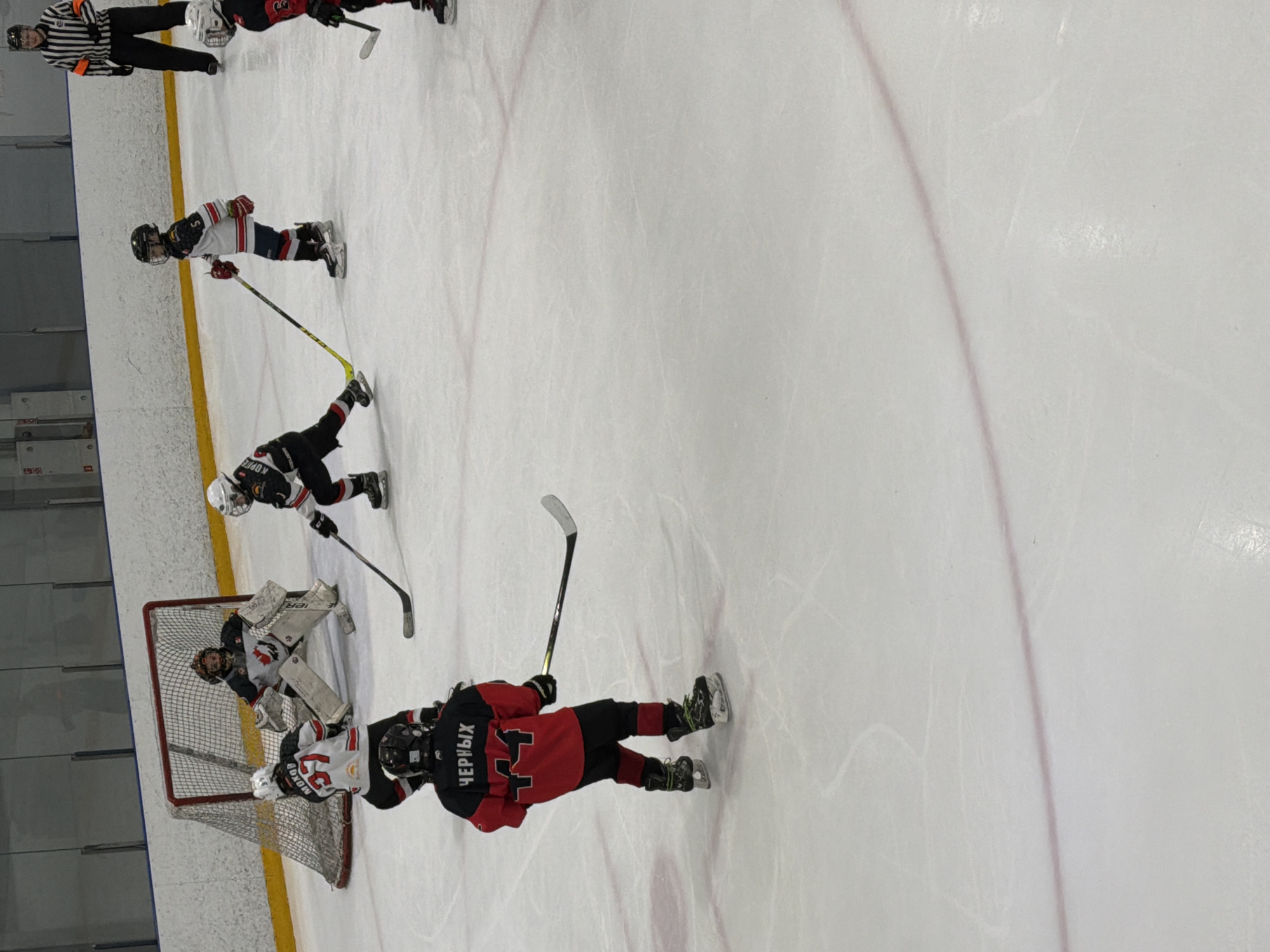 The image shows a youth hockey game in progress on an ice rink. The players, dressed in full hockey gear, are actively engaged in the game near the goal. There's a goalie defending the net. An official in a black and white striped uniform observes the play from the periphery. The rink has yellow boards and red line markings on the ice. The text "ЧЕРНЫХ" is visible on one player's jersey. The city of Votkinsk, Russia is not clearly discernable.