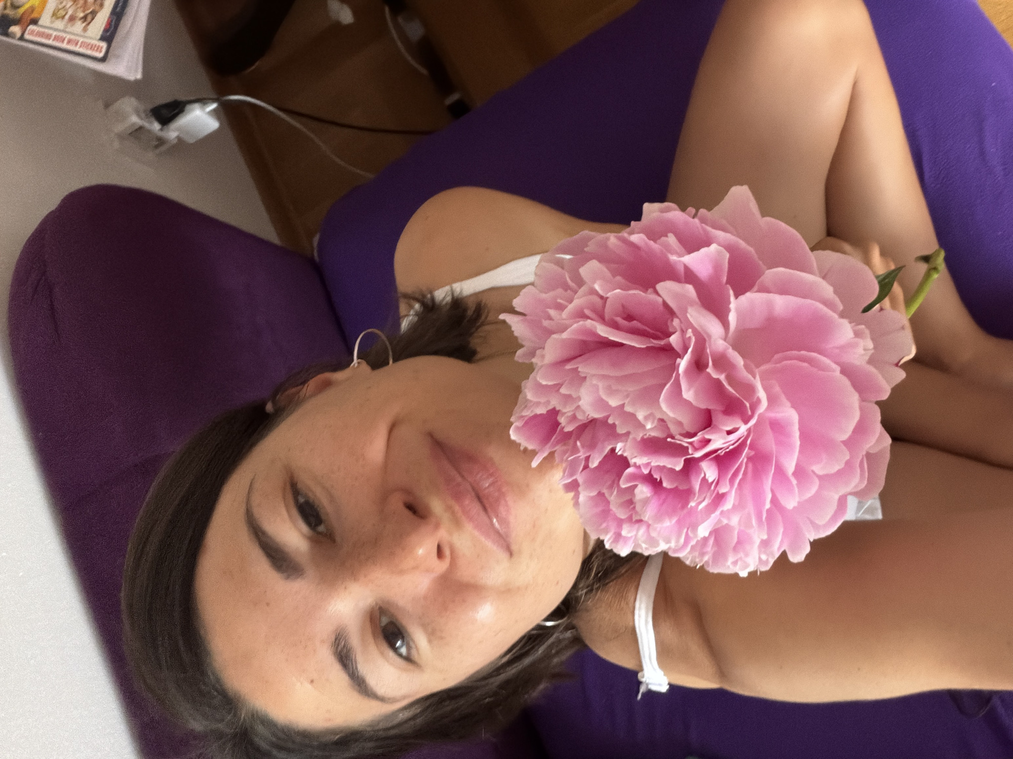 A woman lies on a purple couch, holding a pink peony flower. The background shows a glimpse of a room with a table and a book. The location, Budva, Montenegro, cannot be determined from the visual information.