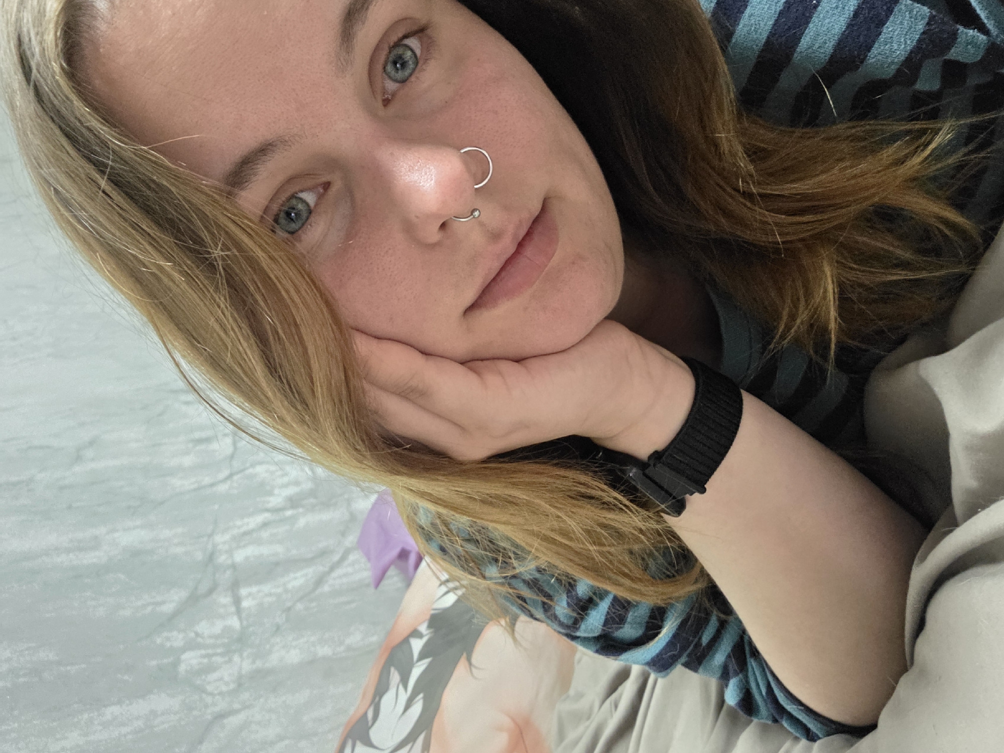 The image shows a young person with blonde hair and blue eyes, lying down and looking at the camera. They are wearing a striped shirt and a watch on their left wrist. The person has a septum piercing with two rings. The background appears to be a bedroom, with a bed and a patterned pillow or blanket visible. The lighting is soft, suggesting it is either indoors or overcast. There are no discernible landmarks or features that would indicate the location within the city of Minsk, Belarus.
