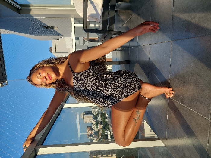 A woman with long, light brown hair is reclining on a balcony overlooking a cityscape. She is wearing a short, patterned dress. Her right arm is extended to the left, resting on the balcony's edge. Her left arm is bent, her hand near her head. Her legs are bent at the knee. An anklet is visible on her right ankle, and a small tattoo is on her lower right leg. Sunlight bathes the scene, casting long shadows on the tile floor. A glass railing surrounds the balcony, with a mesh netting visible above.