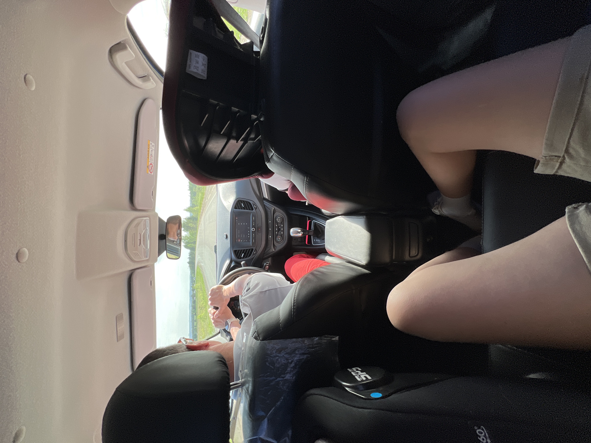 A view from the back seat of a car traveling on a road in Sidorovy Gory, Russia. The driver is visible, wearing a light-colored shirt, red pants, and a watch. Their hands are on the steering wheel. A child car seat is in the back seat, and an individual's leg is visible in the back passenger seat. The car's dashboard, console, and rearview mirror are visible, as well as the external environment seen through the windshield.