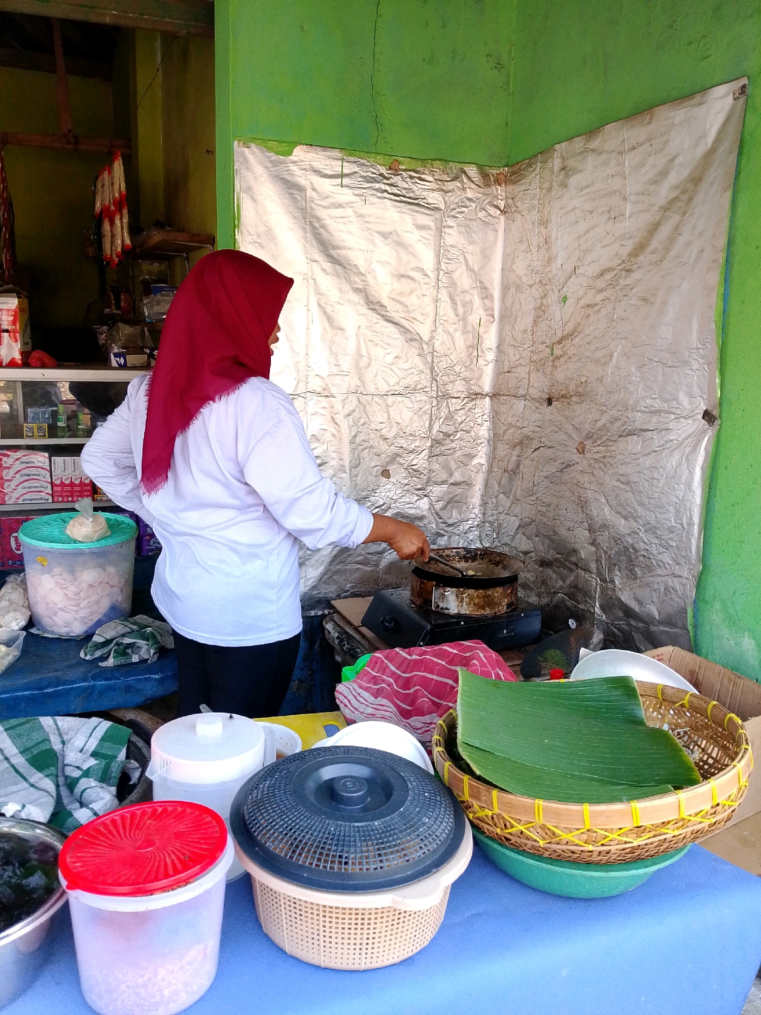 In Banyakan, Indonesia, a woman wearing a red hijab and a white long-sleeved shirt stands in front of a makeshift cooking station. Her right hand is holding a spatula, stirring food in a pan that rests on a portable stove. The cooking area is enclosed by a wrinkled, silvery material, possibly aluminum foil, which covers the walls. The green walls of the room are visible on the sides. In the foreground, several food containers and baskets are arranged on a blue surface. One container, a clear plastic one with a red lid, holds what appears to be small, light-colored food items. Another basket, woven with bamboo, is filled with green banana leaves. A green bowl is placed underneath this basket. To the left, a large, teal plastic container is filled with round, flat, light-colored food items. Shelves behind the woman hold various products, including boxes with red and white packaging. The overall scene depicts a street food vendor preparing food in a modest, outdoor or semi-outdoor setting.