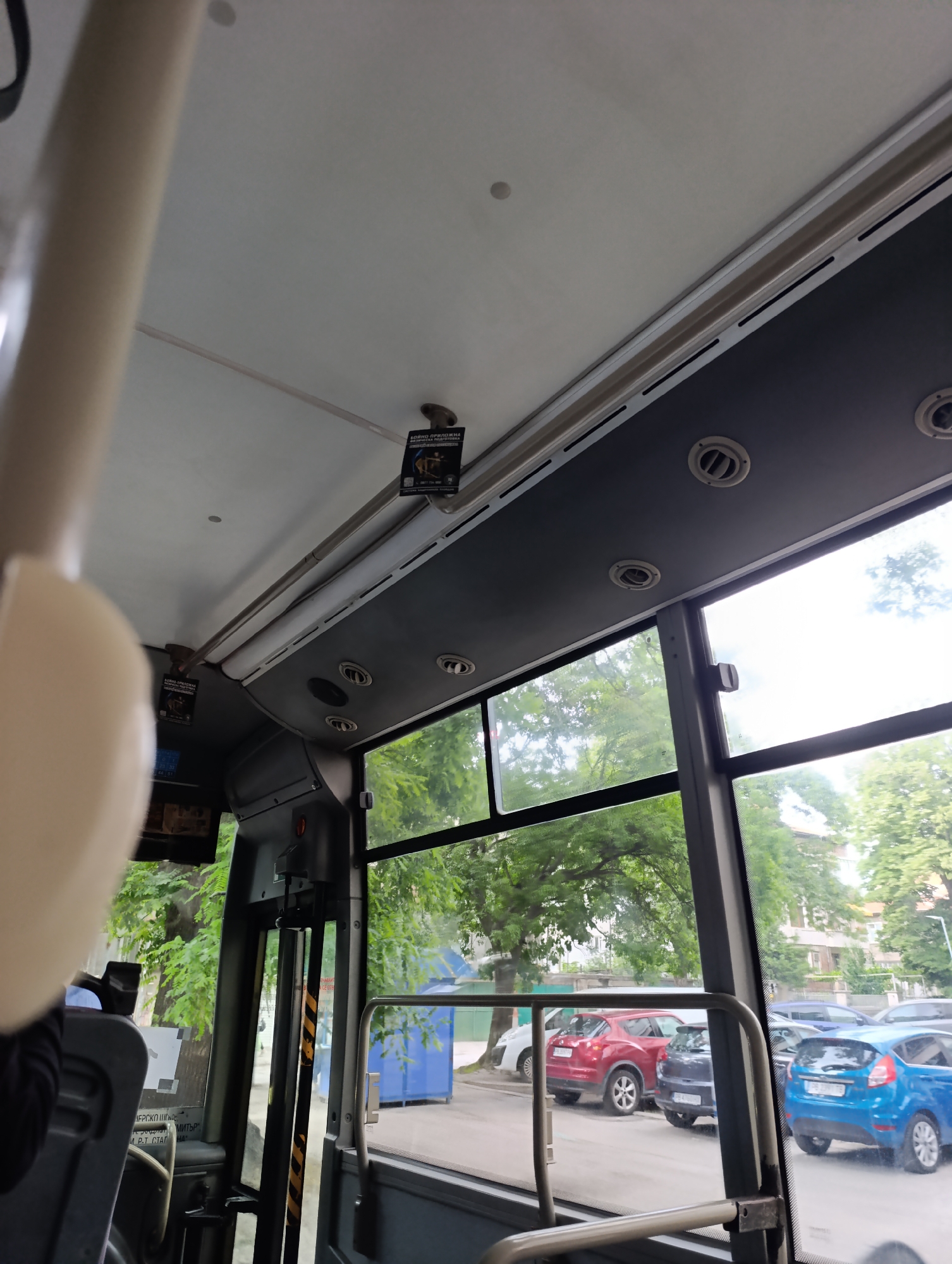 The image captures an interior view from a public transport vehicle, likely a bus, looking towards its right side and front. The perspective is slightly upward, showing the vehicle's ceiling and overhead fixtures.
Inside the vehicle, a light-colored ceiling transitions into a dark gray overhead panel on the right, equipped with several circular air vents. A light-colored, out-of-focus pole or handle is prominent in the immediate foreground on the left. Two small, black rectangular signs are affixed to the ceiling structure. The visible text on these signs, in Bulgarian, reads "БОЙНО ПРИЛОЖНА СТРЕЛБА ЗА МЛАДЕЖИ И ВЪЗРАСТНИ" (COMBAT APPLIED SHOOTING FOR YOUTH AND ADULTS), followed by a phone number "0897 704 980". A metal handrail runs horizontally along the bottom of the large window, and a vertical bar with yellow and black hazard stripes has partially visible text "ВНИМАНИЕ" (ATTENTION/WARNING).
Through the bus windows, an urban street scene is visible. Several cars are parked along the curb, including a red SUV, a dark gray hatchback, a bright blue Ford Fiesta, and part of a white car. The blue car's license plate begins with "PB", a registration code for Plovdiv, Bulgaria, confirming the location provided. Lush green trees line the street, suggesting either late spring or summer. Buildings are visible behind the foliage, and a blue dumpster or container is present on the sidewalk. The lighting suggests it is daytime, with soft, possibly overcast light. No people are clearly visible. The overall scene is calm, depicting an ordinary moment of transit.