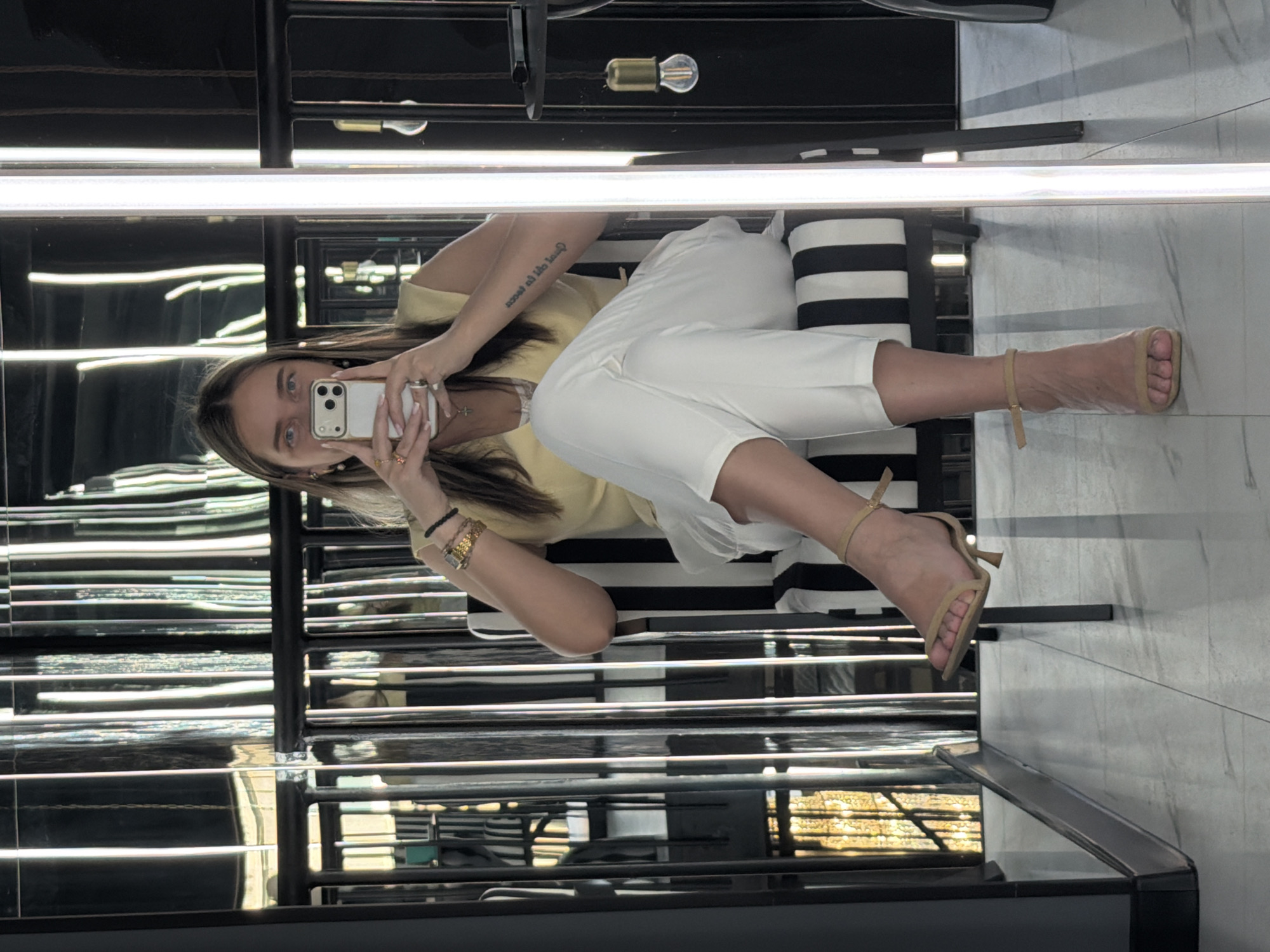 A woman is reflected in a mirror. She sits on a black and white striped seat, wearing a yellow top, white pants, and tan heeled shoes. She is holding a smartphone and looking at her reflection. The reflection shows a dark background with horizontal lines, possibly blinds or shelves. The location is unknown and there is no evidence to suggest it is Moscow, Russia.