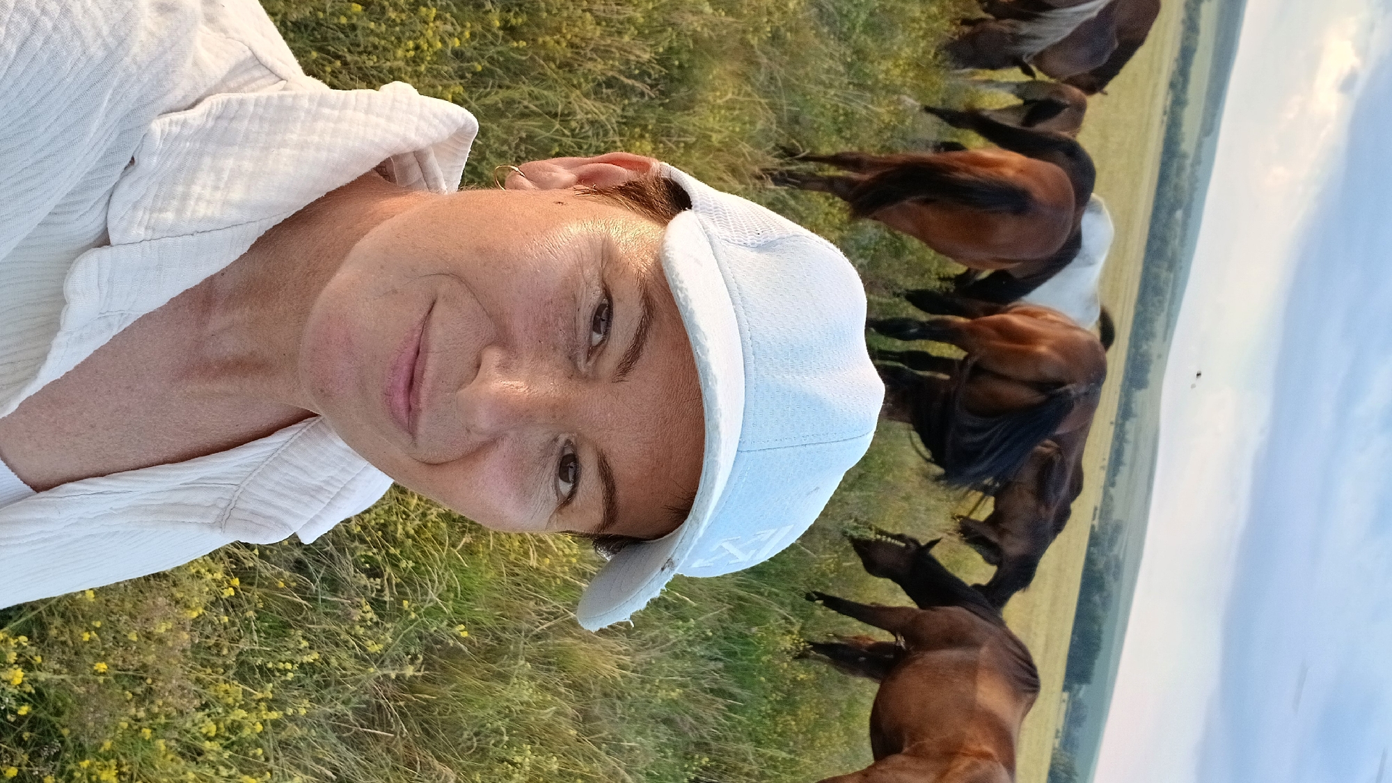 A woman wearing a white cap and white collared shirt takes a selfie in a grassy field with a herd of horses in the background. The sky is cloudy with hints of sunset colors. The location is Nadezhdenka, Russia.