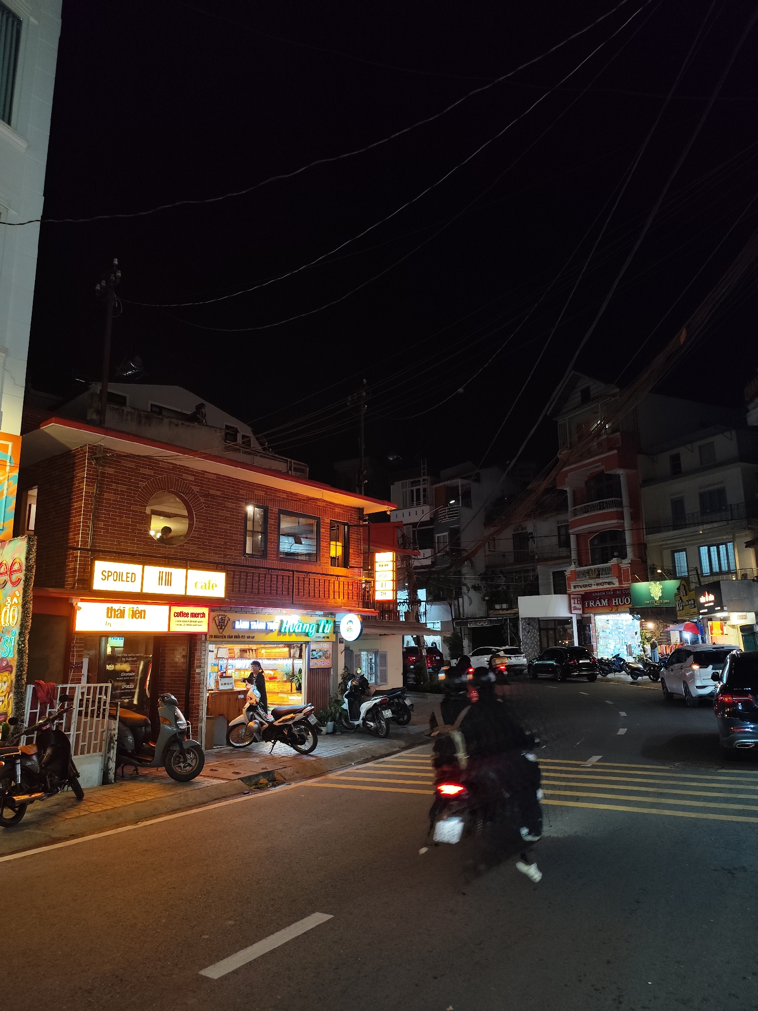 The image shows a street scene in Da Lat, Vietnam, at night. The foreground features a street with yellow and white lane markings, with a motorbike moving towards the viewer. Parked motorbikes are along the sidewalk on the left side of the road. Several businesses are visible on the left, including a cafe called "SPOILED," identified by its sign, and a shop named "Thai Lien," and "Hoang Tu". The buildings are brick or painted in bright colors, with illuminated signs. In the background, other buildings and businesses can be seen along the street. The lighting indicates it is night, with streetlights and business signs illuminating the scene. People are visible in the storefronts. The overhead is filled with a dense network of power lines. Cars are parked along the right side of the street.