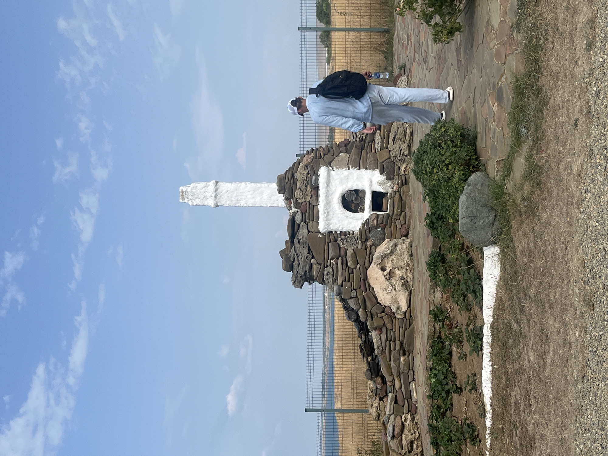 The image depicts an outdoor scene in Taman', Russia. In the foreground, a person wearing a light blue long-sleeved shirt, light blue pants, a white cap, and a backpack walks away from the camera on a stone-paved path. The person is holding a water bottle in their right hand.
The central focus is a stone structure resembling a primitive oven or furnace, topped with a tall, white, cylindrical chimney. The structure is made of various natural stones and has a white, recessed opening. Green bushes and dry grass surround the base of the structure and the path. A metal fence runs horizontally behind the structure, separating it from a field of dry, golden grass and a body of water in the distance. The sky above is blue with scattered white clouds. The overall impression is a rustic, outdoor monument or landmark.