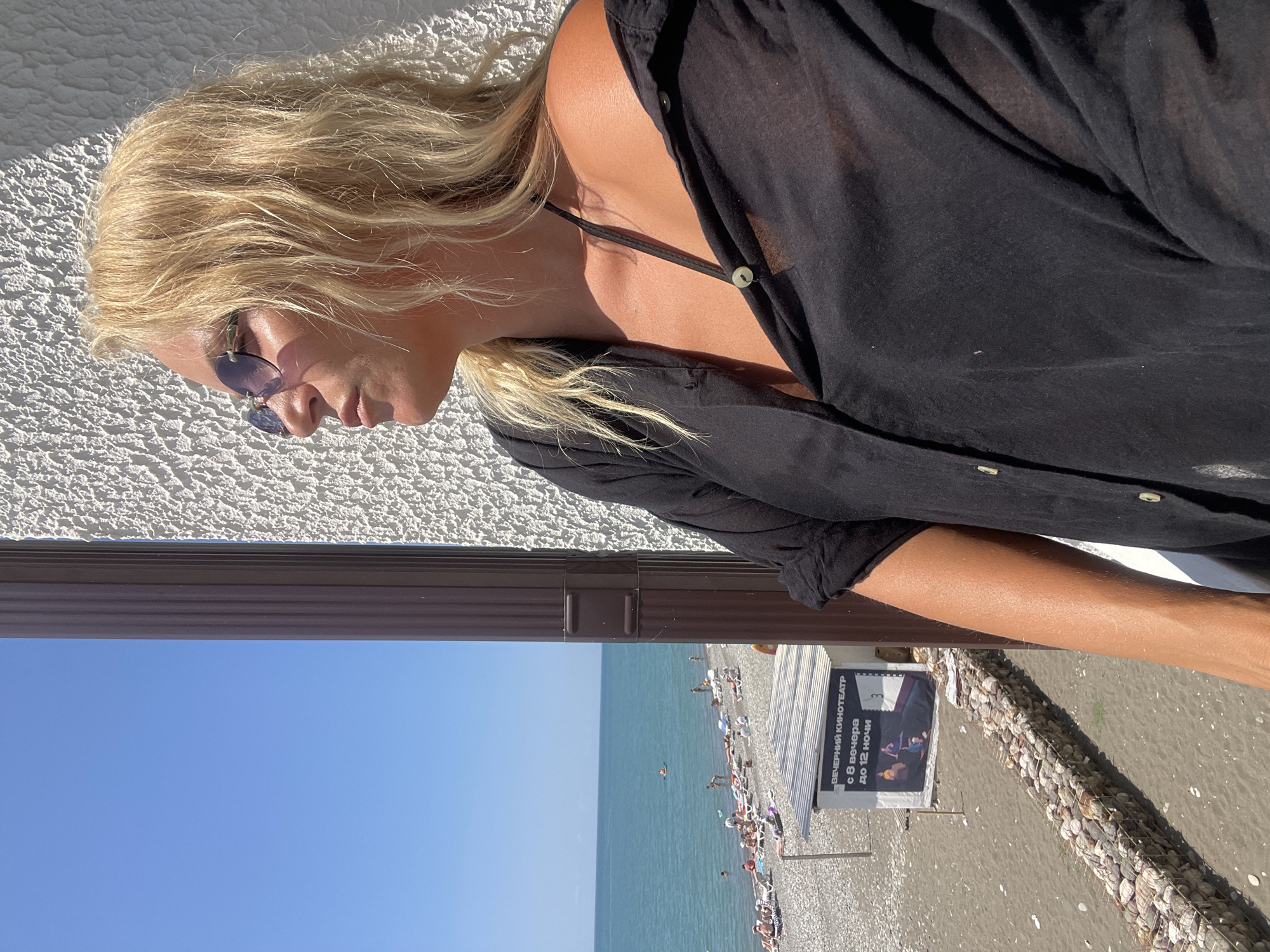 A woman with blonde hair and sunglasses stands on a balcony, looking out at a beach and the sea. She is wearing a black open shirt and a dark bikini top. The beach is populated with people sunbathing and swimming in the clear blue water. A sign for an "Evening Cinema" is visible on the beach. The scene is brightly lit by the sun. The location is Giachrypsh, Abkhazia.
