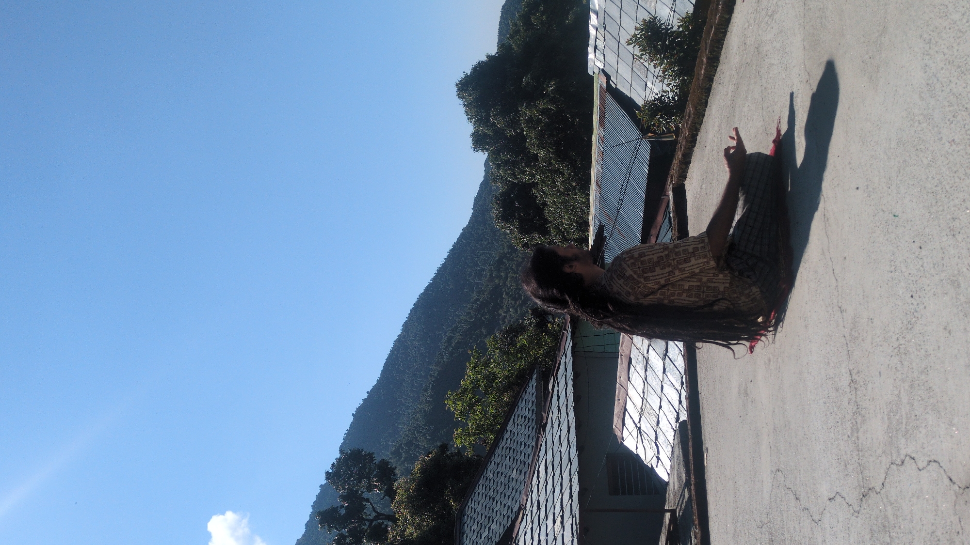 A man with long dreadlocks is sitting on a rooftop in Bhagsu, India. He is positioned on the right side of the frame, with his back toward a metal fence, casting a shadow on the concrete rooftop. He is wearing a patterned shirt and plaid pants. In the background, a mountain covered in dense green trees extends towards the clear blue sky on the left side of the frame. The rooftop appears to be attached to a building with a tiled roof that is partially visible.