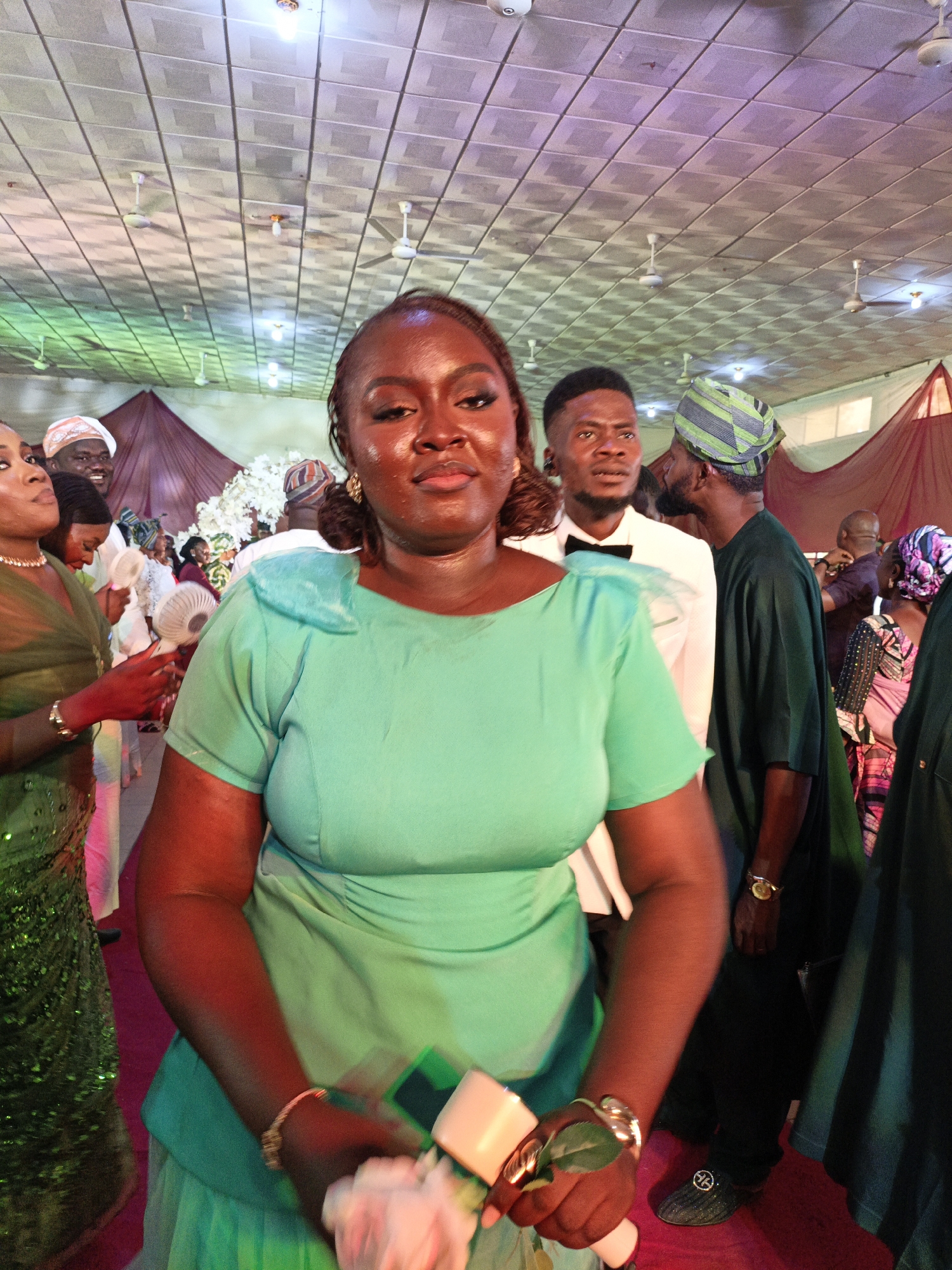 The image, likely taken in Fagge D2, Nigeria, shows an indoor wedding reception. A woman in a teal dress is in the foreground, holding what appears to be a corsage or bouquet. Behind her are other attendees. One man wears a white suit with a black bow tie. Another wears a traditional Nigerian hat. The setting appears to be a banquet hall, indicated by the ceiling with multiple ceiling fans and draped fabrics.
