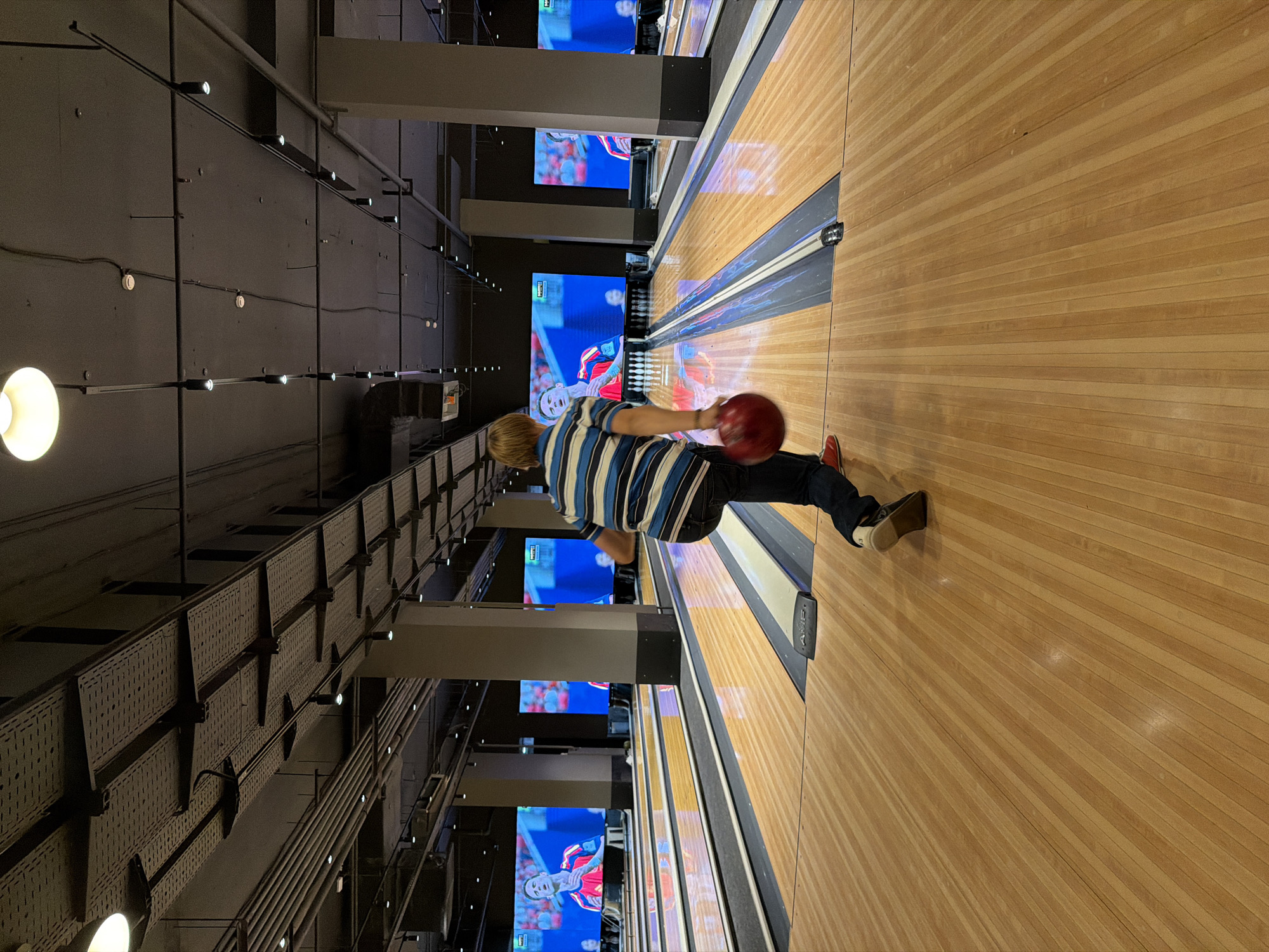 The image shows a young person with blond hair, wearing a blue and white striped shirt and dark jeans, bowling in an indoor bowling alley. They are in the process of throwing a red bowling ball down a polished wooden lane. In the background, large screens display images of a soccer player. The bowling alley has multiple lanes, each marked with dark and light wood. The ceiling is industrial, with visible pipes and lighting fixtures. The overall environment suggests a modern entertainment venue. The location of the bowling alley in Izhevsk, Russia, cannot be determined from the image itself, as there are no discernible landmarks or identifying features.