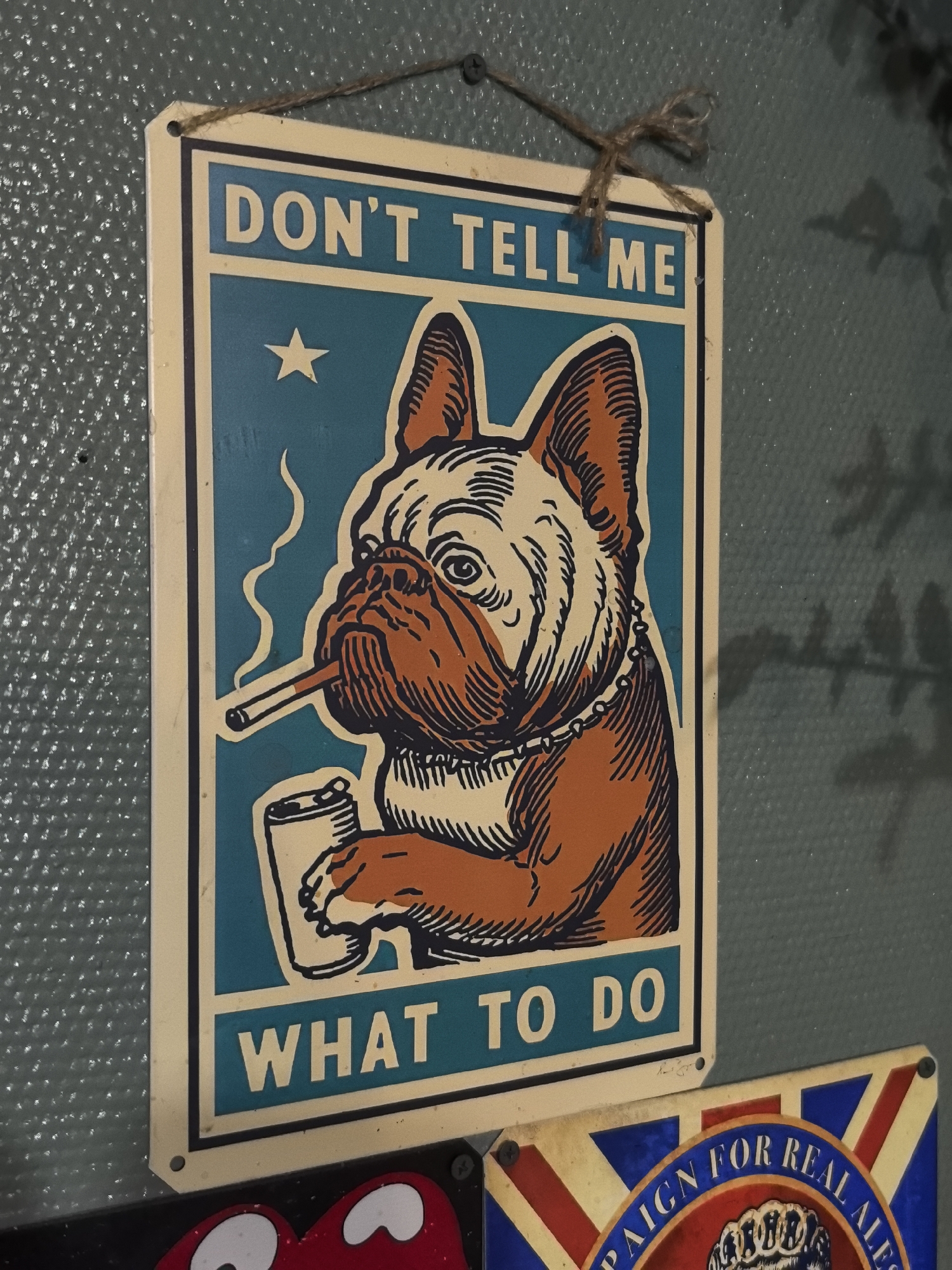 A close-up shot depicts a rectangular metal sign hanging on a wall. The sign features a drawing of a dog, possibly a bulldog, smoking a cigarette and holding a beverage can. The background of the sign is teal with a white star near the upper left corner. Bold white letters read "DON'T TELL ME" at the top of the sign and "WHAT TO DO" at the bottom. The edges of the sign are beige.
Below the sign, partially visible, is another sign featuring the Union Jack flag and the words "PAIGN FOR REAL ALE".
The scene appears to be indoors, likely in a pub or similar establishment, given the subject matter of the signs. The backdrop is a textured gray-green wall. The photo was taken with a shallow depth of field.