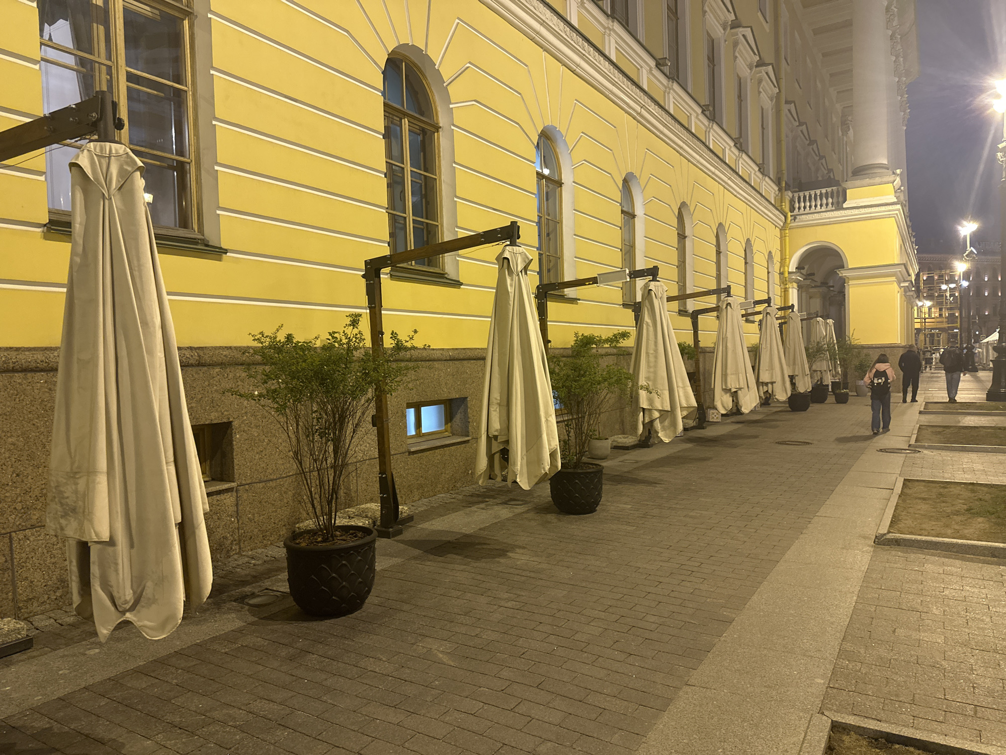The image depicts a street scene in Saint Petersburg, Russia, at night. Along a yellow building with arched windows, closed white umbrellas are suspended from metal frames, alternating with potted plants. The sidewalk is paved with brick. A few pedestrians are walking in the distance. Streetlights illuminate the area.