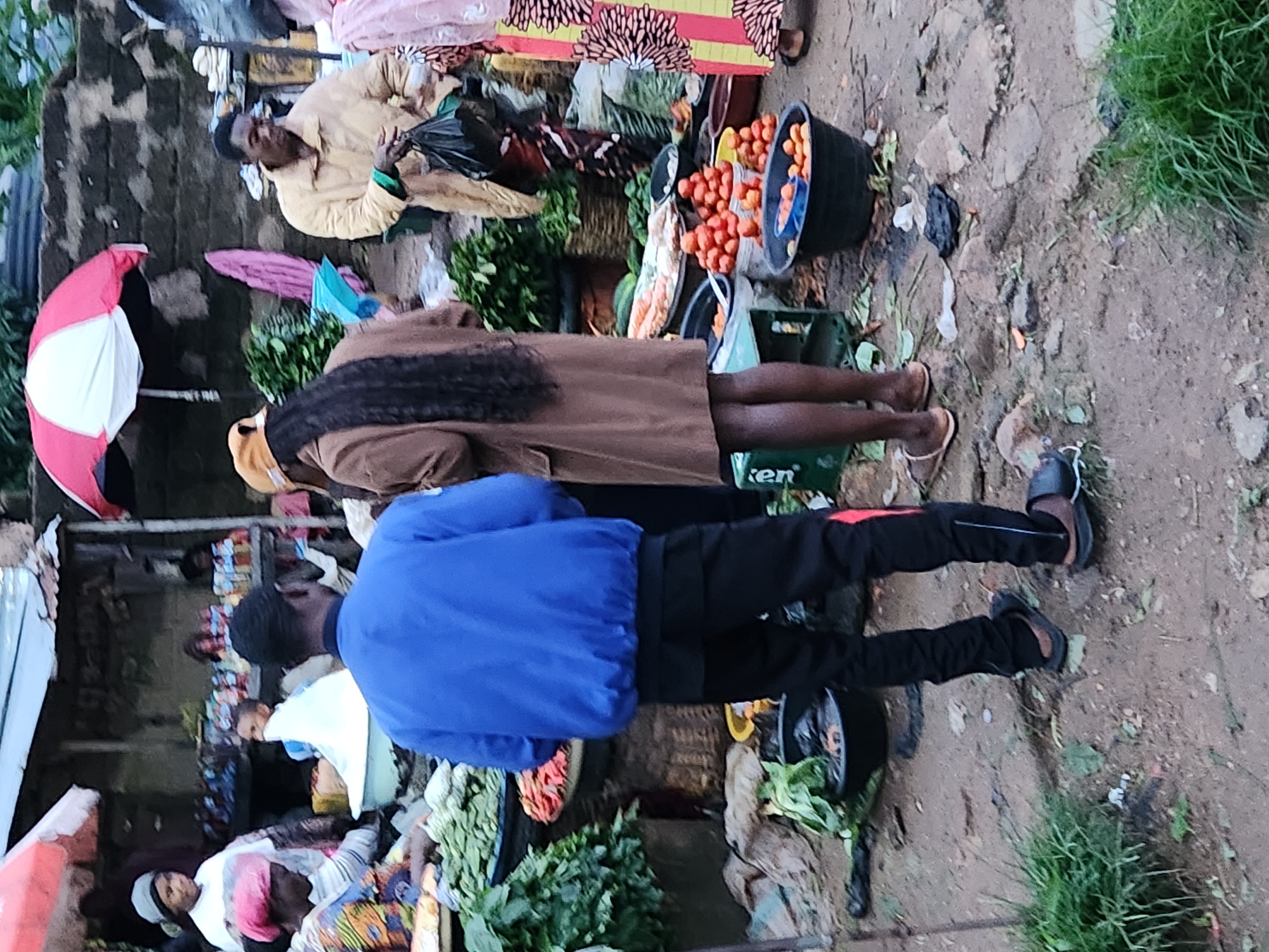 The image depicts an outdoor market scene in Kabong, Nigeria. In the foreground, a person wearing a blue jacket and black pants stands facing away from the camera. To their right, a person in a brown coat with long dark hair also stands facing away. The background shows stalls with various goods, including produce like tomatoes and leafy greens. Several individuals are visible in the background, some appearing to be vendors and shoppers. The market appears to be set up on a dirt or unpaved ground. The overall impression is a busy marketplace with a variety of people and goods on display.