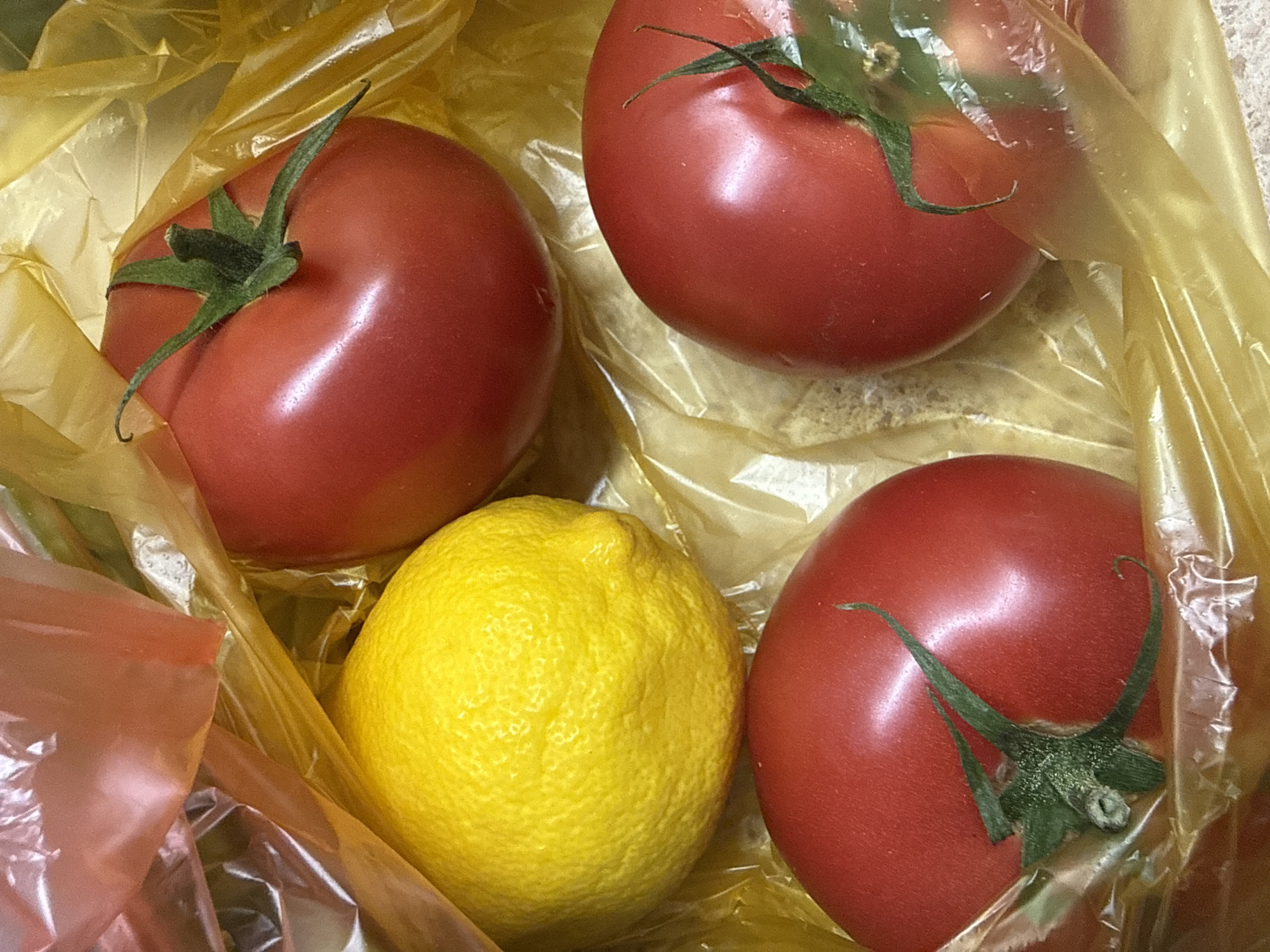 A high-angle, close-up shot shows three ripe red tomatoes and one bright yellow lemon nestled inside a translucent yellow plastic bag. The tomatoes are round and smooth with green stems and leafy calyxes. The lemon is also round and textured, with a slightly bumpy rind. The plastic bag is crinkled and reflects light, obscuring parts of the produce. The background is out of focus, suggesting a shallow depth of field.