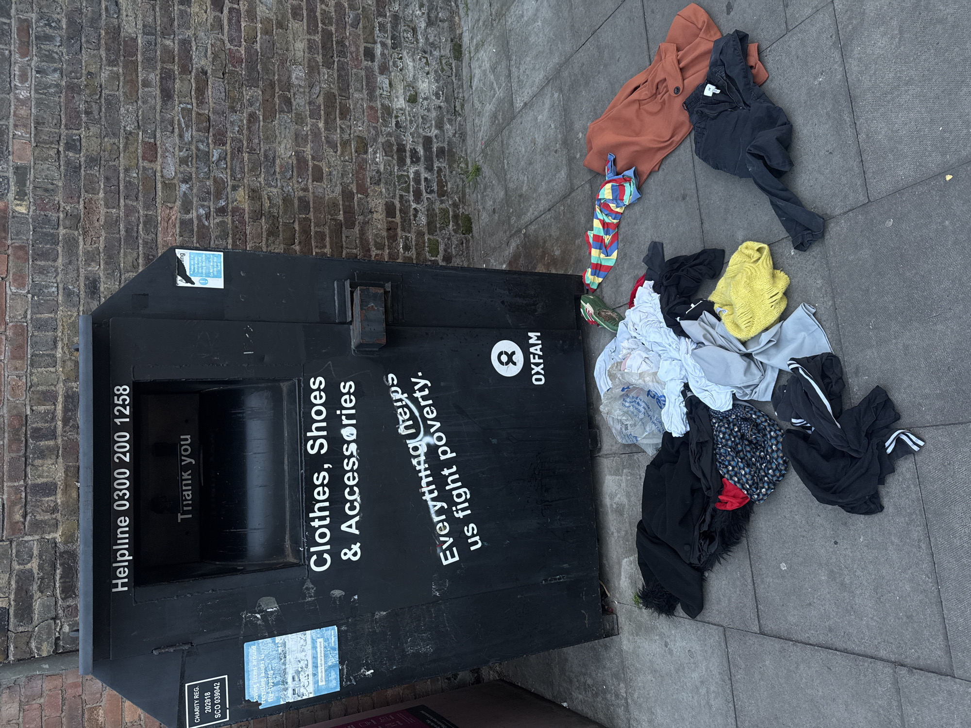 An overhead outdoor shot displays a black Oxfam clothing donation bin on a grey paved sidewalk, next to a weathered brick wall. The bin, oriented vertically in the left part of the frame, features white lettering that reads "Helpline 0300 200 1258," "Clothes, Shoes & Accessories," and "Everything helps us fight poverty." The Oxfam logo and name are visible on its side, and "Thank you" is printed inside the donation slot. Small stickers on the bin include a charity registration number ("CHARITY REG. 202918 SCO 039042") and a warning stating, "Leaving items around recycling banks is fly-tipping. Offenders will be prosecuted."
A significant pile of assorted clothing and textiles is strewn across the sidewalk to the right of the bin, extending into the foreground. The garments include an orange long-sleeved top, black trousers, a vibrant multi-colored striped top, a ripped yellow sweater, and various crumpled white, grey, and black items. A clear plastic bag filled with white fabric is also visible among the pile.
The scene takes place during daytime, under what appears to be overcast conditions, as suggested by the even lighting and lack of harsh shadows. The setting is an urban environment, characteristic of London, United Kingdom, with the brick wall providing a common local architectural element. No people are visible in the image, leaving the focus on the donation bin and the discarded clothes.