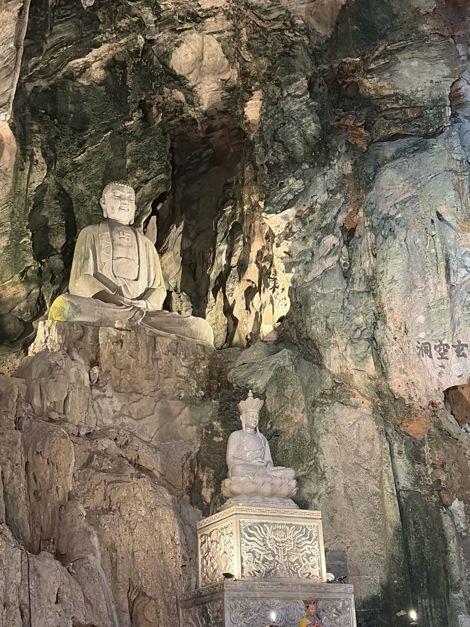 The image depicts the interior of a cave, likely a religious site, featuring two Buddha statues carved from stone. The larger statue, on the left, is seated cross-legged on a rocky base, with its hands resting in its lap. It wears a serene expression and is adorned with a robe. To the right and slightly lower is a smaller seated Buddha statue, also cross-legged, wearing a crown. This statue is placed on an ornate, multi-tiered pedestal adorned with intricate carvings. The cave walls are rough and uneven, with layers of rock visible, suggesting a natural geological formation. Lighting, possibly artificial, illuminates the statues and the cave environment, casting shadows and highlighting the textures of the rock. Chinese characters are inscribed on the rock face to the right of the larger statue. The presence of these statues and the cave setting suggest this is a temple or a place of worship. This scene is located within the Marble Mountains, specifically in the Huyen Khong Cave, in the city of Đà Nẵng, Vietnam.