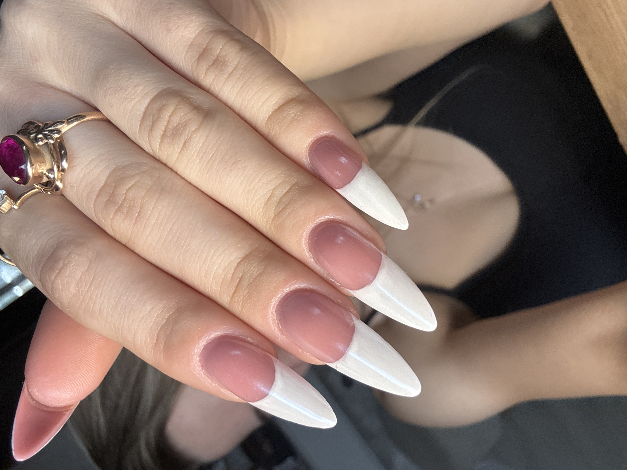 A close-up photograph features a woman's hand adorned with a vintage gold ring set with a deep red stone. The ring is on the ring finger. Her fingernails are long, stiletto-shaped, and painted in a modern French manicure style, with a pale pink base and bright white tips. The hand is positioned prominently in the foreground. The background shows a blurry outline of a black garment and hints of a necklace. The scene appears to be indoors, but the specific location in Noviy, Russia cannot be determined from the image.