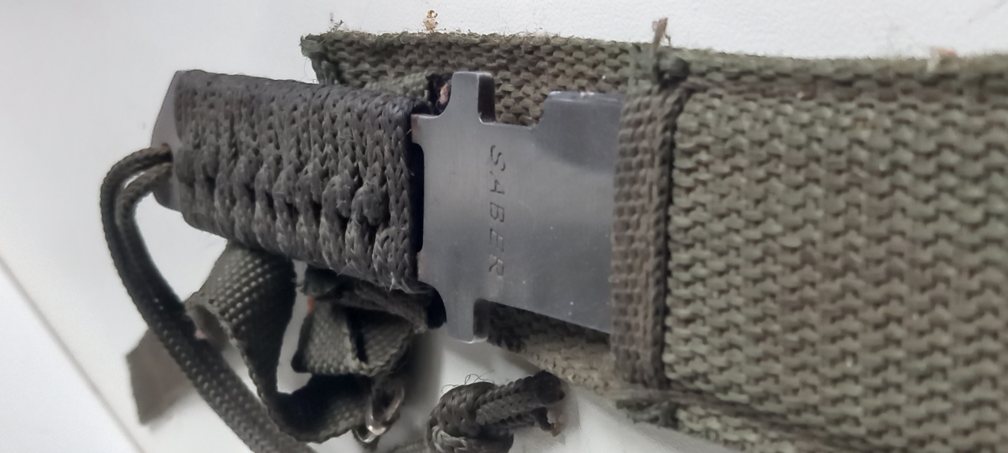 A close-up shot captures a military-style or outdoor knife partially sheathed in an olive green fabric. The knife features a dark, intricately woven handle, possibly made of paracord, which extends to the left of the frame. The blade, dark gray or black and metallic, is partially visible, emerging from the fabric sheath. Vertically inscribed on the visible portion of the blade is the word "SABER."
The sheath itself is constructed from a robust, textured, woven fabric, likely canvas or nylon webbing, and appears well-worn. Various dark-colored straps and cordage are visible around the handle and base of the sheath, suggesting a carrying system. The item is resting against a plain, light-colored background, possibly a wall or table, which is out of focus, emphasizing the knife and sheath. There are no people visible, and no specific activity is taking place; the knife appears to be stored or static. The overall impression is of practical, rugged gear. The location context is Kryvyi Rih, Ukraine.