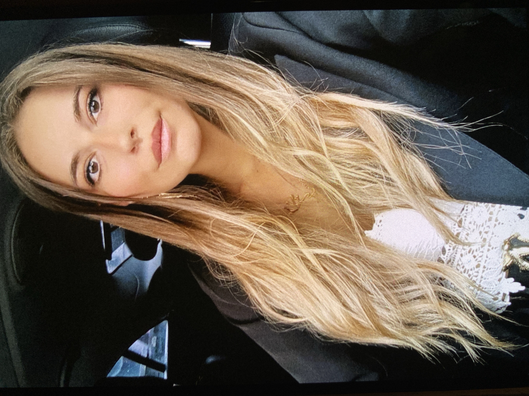 A young woman with long, wavy, light brown and blonde hair is captured in a tilted, close-up portrait, likely a selfie. She has a fair complexion, warm brown eyes, and light pink lips, showing a faint, pleasant smile. She is wearing a dark-colored jacket or coat, beneath which a white top with intricate lace or crochet details is visible on her right side. A delicate gold-colored necklace can be seen around her neck. The background is dark and appears to be the interior of a vehicle, possibly a car, with dark, textured upholstery. No specific time of day or weather conditions are discernible from the image. The photo's location context is Bankya, Bulgaria. No text is visible in the media file.