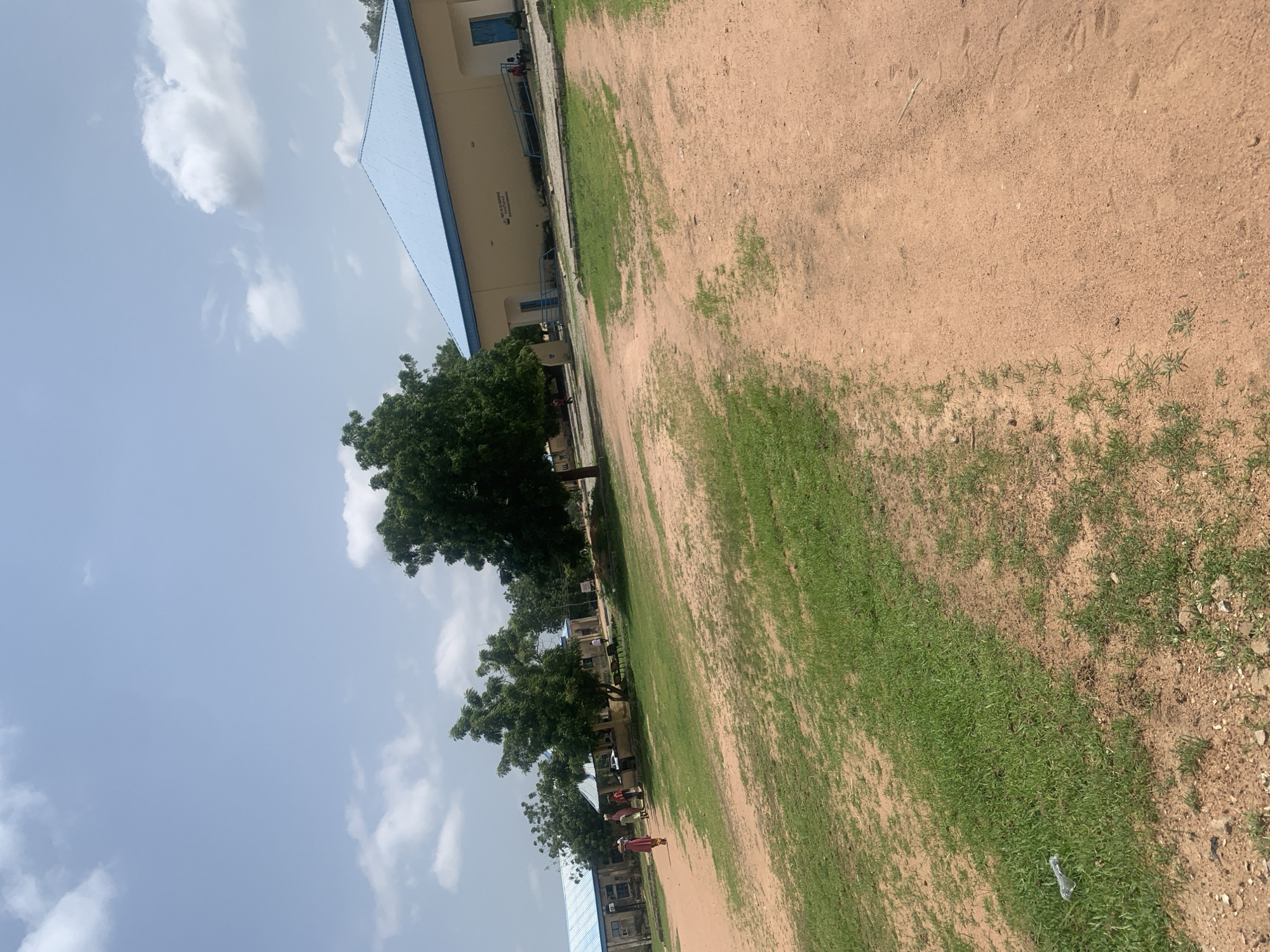 A group of people are seen walking on a dirt path in front of a building with a blue roof. The building is light yellow and appears to be a school or government building. There are several trees in the foreground and background. The sky is blue with some clouds. The location is the Taraba State University, Jalingo, Nigeria.