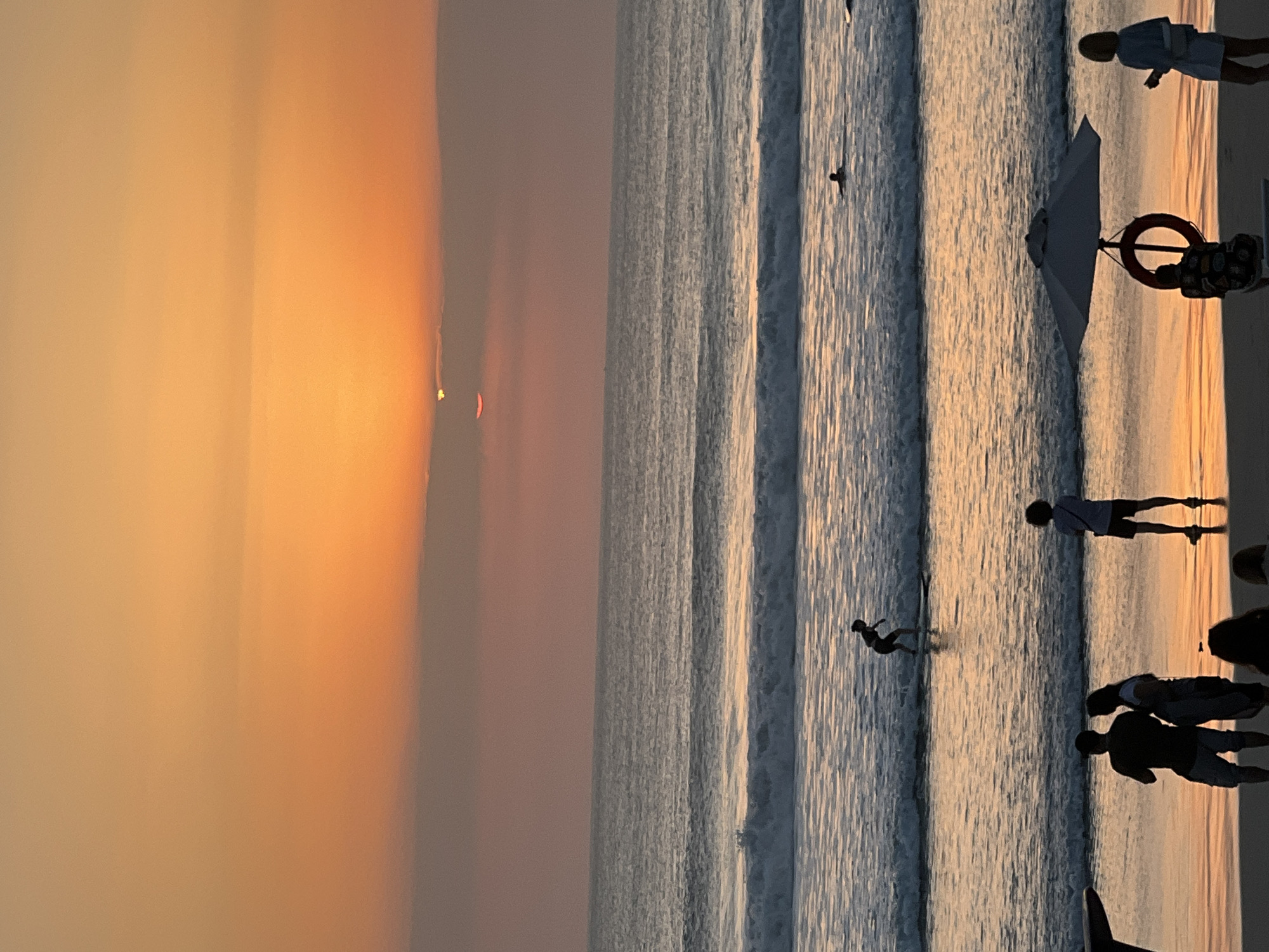 This is a vertical, eye-level photograph of a beach scene in Kuta, Indonesia during sunset. The sun is low on the horizon, partially obscured by clouds, casting an orange glow across the sky and reflecting on the water. The ocean has multiple sets of waves rolling towards the shore. In the middle ground, a lone surfer is riding a wave. Further out, another person is visible in the water, possibly swimming. In the foreground, several people are gathered on the sand, silhouetted against the sunset. Some are standing in groups, while others are sitting. A beach umbrella and a life preserver are also visible on the sand. The overall impression is one of a serene and beautiful evening at the beach.