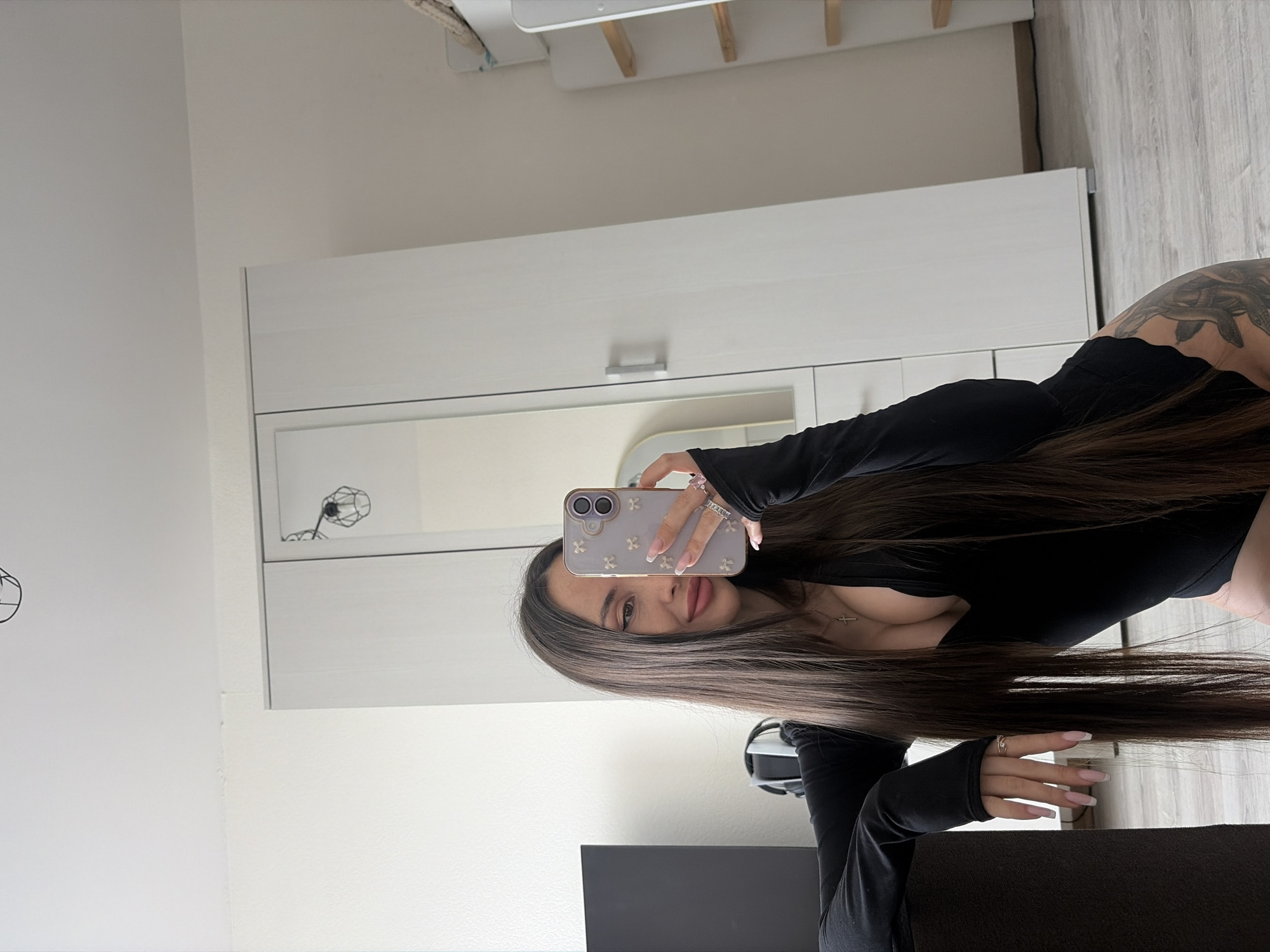 A woman with long brown hair, a tattoo on her upper back, and a black long-sleeved top is taking a selfie with a phone. She is indoors, likely in a room with a wardrobe. The location is not discernible as Saint Petersburg, Russia.