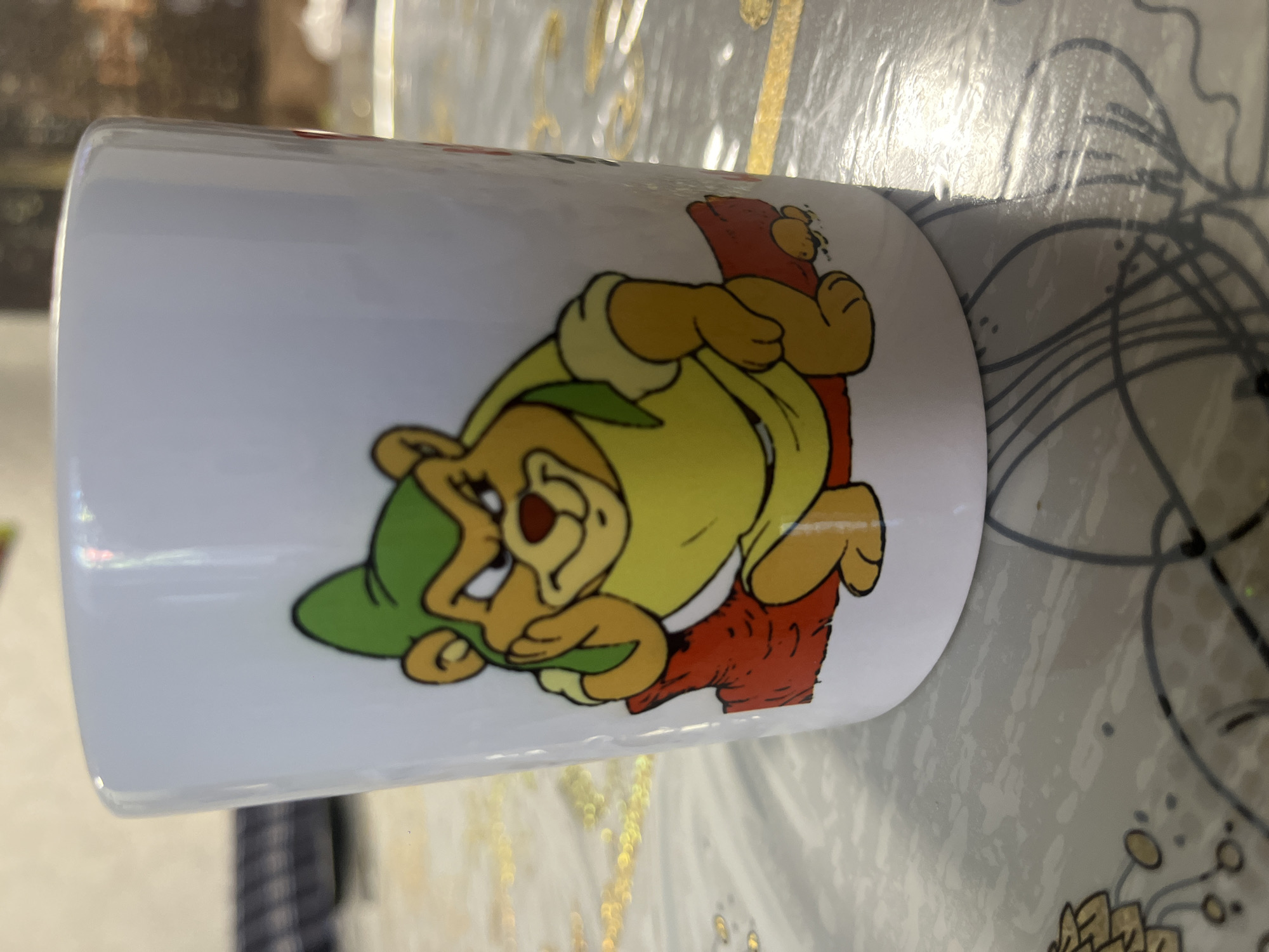 A white ceramic mug with a cartoon bear illustration is placed on a patterned surface. The bear is depicted wearing a yellow tunic and a green cap, with its left arm resting on a red log. The background of the mug is white. The surface below the mug has a light gray base with darker gray lines and circular patterns. The lighting suggests an indoor environment. The image does not contain any information about the city of Vladimirovka, Russia.