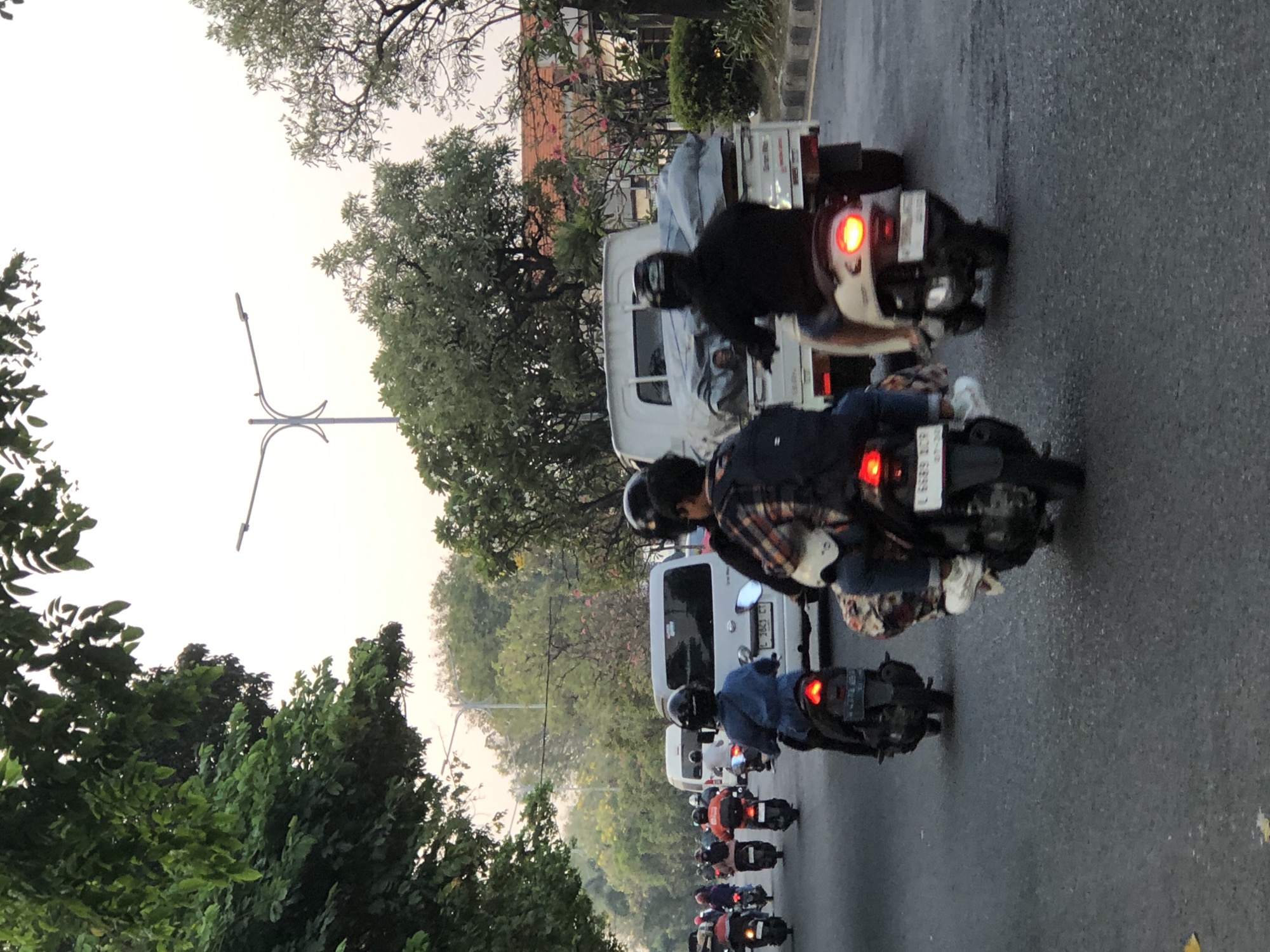 The image depicts a street scene in Sumorame, Indonesia, likely during the late afternoon or early evening, with a traffic jam of motorcycles and a few white vans. The road is asphalt, and trees line the sides of the street. In the foreground, several motorcycles are visible, with riders wearing helmets. One motorcycle in the center has a rider with a patterned shirt and a backpack, and another individual is seated behind them. To the right, a motorcycle carries a rider with a light-colored jacket and a white helmet. Further ahead, a white van with a covered load is stuck in traffic, with another white van positioned behind it. In the distance, more motorcycles and vehicles are seen, suggesting a significant traffic congestion. A street light stands tall on the right side of the road, and a few buildings are visible in the background through the trees. The sky is overcast with a soft, diffused light.