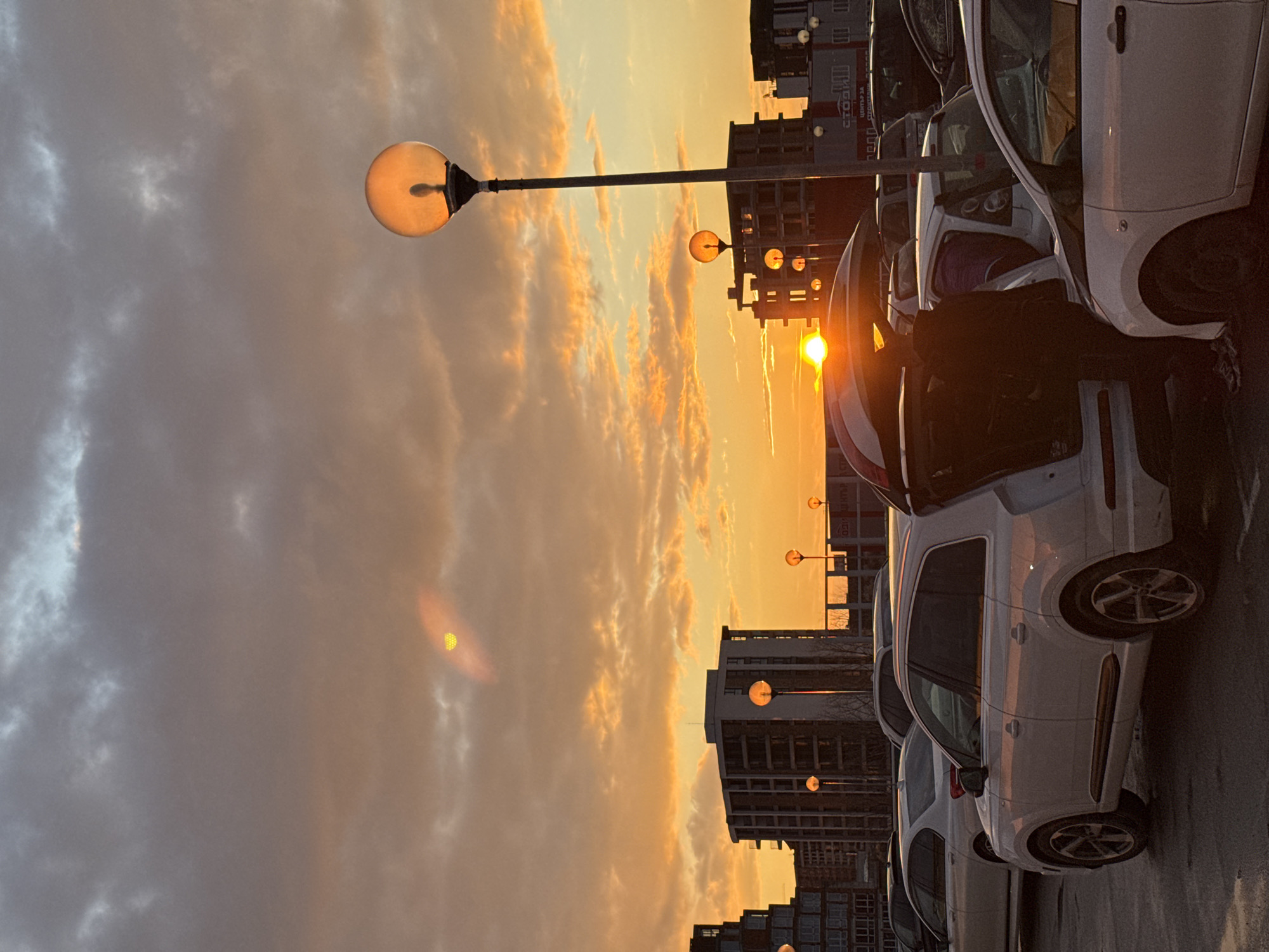 The image depicts a row of white cars parked on a street in Burgas, Bulgaria. In the background, buildings are silhouetted against a sunset sky with orange and gray clouds. Several streetlights with round globes are visible, some illuminated. A person is partially obscured by a car. The angle of the image suggests it was taken from a low vantage point.