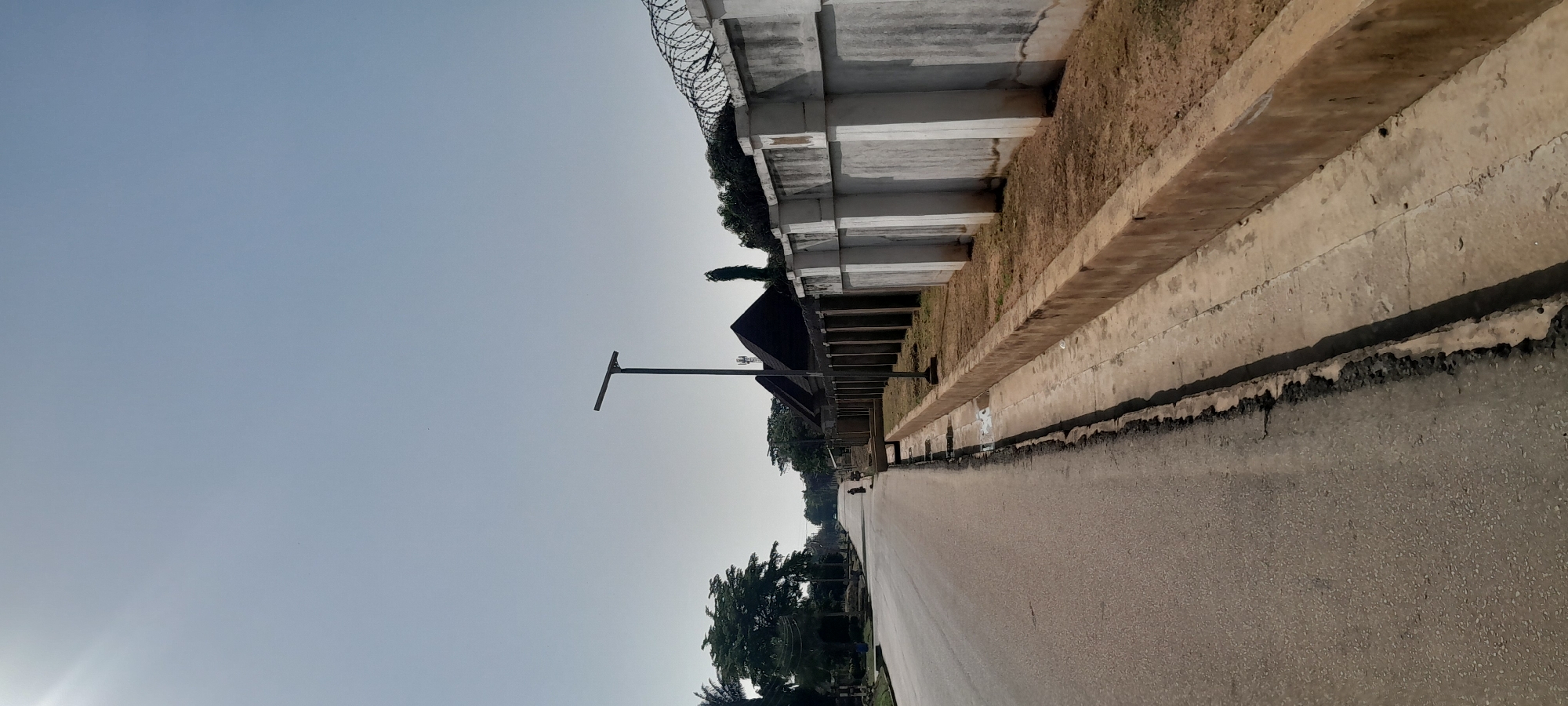 The image captures a wide, paved road in Jalingo, Nigeria, under a clear, pale blue sky. The road curves slightly to the left and recedes into the distance. On the right side of the road, there is a concrete curb and a narrow drainage channel. Beyond the channel, a substantial concrete wall runs parallel to the road, topped with barbed wire coils. The wall is punctuated by large, rectangular white pillars. Behind the wall, glimpses of greenery and dark-roofed structures, possibly houses or commercial buildings, are visible. To the left of the road, trees and vegetation line the side, and a lamppost with a solar panel is positioned near the middle of the frame. A lone person on what appears to be a motorcycle is visible in the distance on the road. The overall impression is of a well-maintained, possibly suburban or semi-commercial area on a bright, clear day.