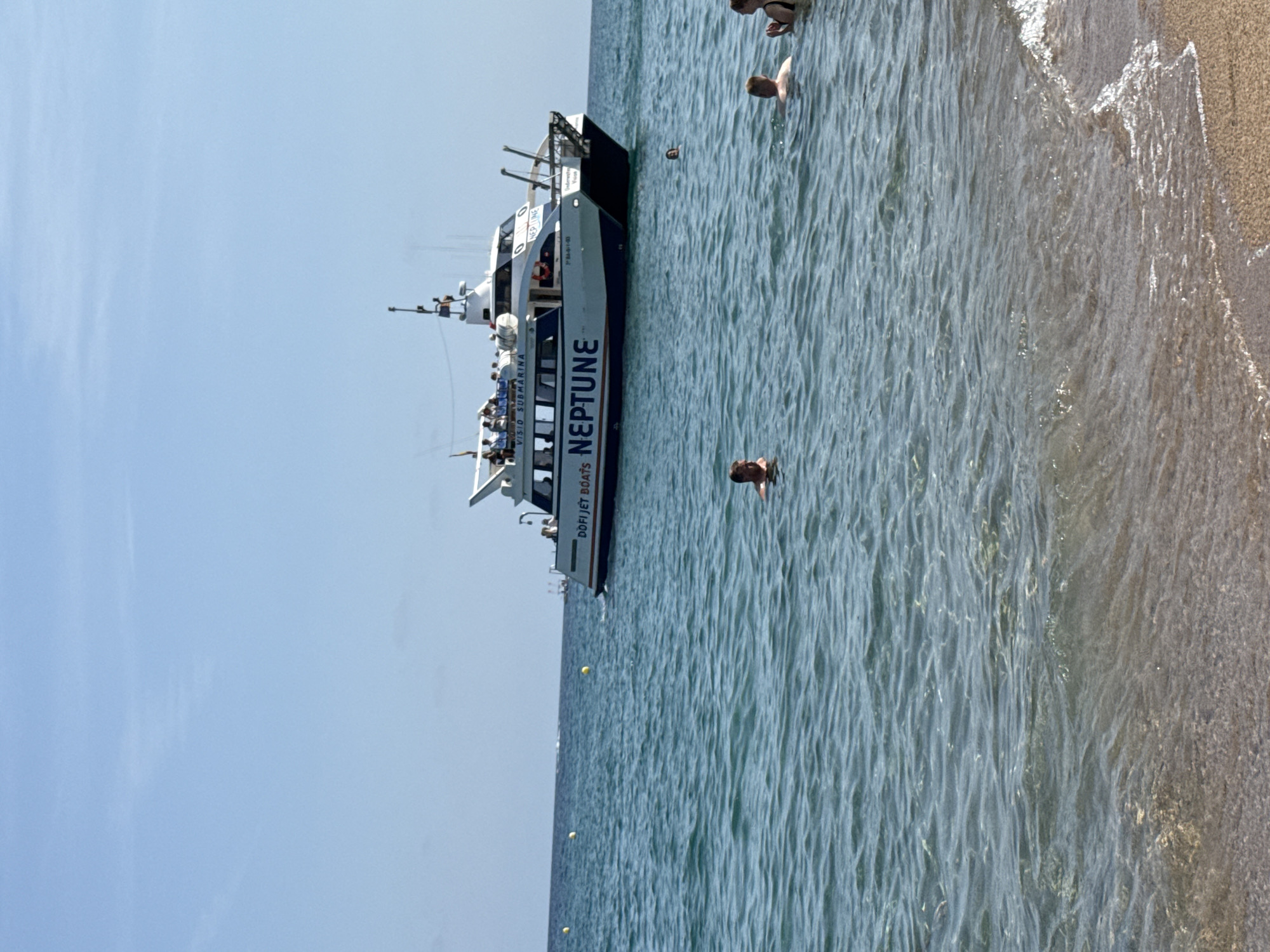 The image depicts a daytime scene at the beach in Blanes, Spain. In the foreground, the shallow, clear water of the Mediterranean Sea gently laps onto a sandy shore. Several individuals are visible swimming and wading in the water.
Further out in the sea, a white catamaran boat named "NEPTUNE" is cruising. The boat is identified as a "VISIO SUBMARINA" vessel, suggesting it offers underwater viewing tours. The boat is white with blue trim and has multiple decks, with passengers visible on the upper deck. The text "DOFI JET BOATS" is also visible on the side of the boat.
The sky is clear and blue, indicating good weather conditions. The overall scene conveys a typical summer day at a coastal resort town.