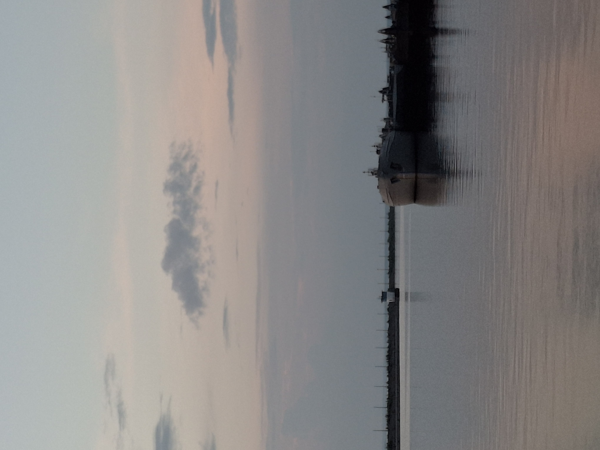 The image captures a serene waterfront scene at dusk in Chaikovsky, Russia. The sky is painted with soft hues of pink and gray, with a few wispy clouds. In the foreground, a calm body of water reflects the muted colors of the sky. Along the horizon, a long, low breakwater stretches across the scene, punctuated by a small, white structure resembling a lighthouse or control tower. To the right of the breakwater, a cluster of dark, unlit vessels are moored. The closest vessel, a light-colored ship with a rounded bow, is prominently featured in the mid-ground, its hull partially submerged and casting a subtle reflection on the water. The overall impression is one of quiet stillness, characteristic of a port at the end of the day.