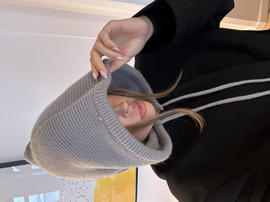 An individual poses indoors within the city of Unknown, Unknown. The subject wears a light grey ribbed knitted hood, pulled forward to obscure the upper face. Below the hood, a black garment with light grey drawstrings is visible. The lower portion of the subject's face, including glossy, full lips and strands of light brown hair, is exposed. A right hand, with light pink manicured fingernails, is positioned at the rim of the hood, appearing to adjust or hold it. The background consists of a light-colored wall. To the left, a portion of an artwork featuring yellow, white, and small black elements is discernible. A faint vertical line, possibly a door frame, is present on the right.