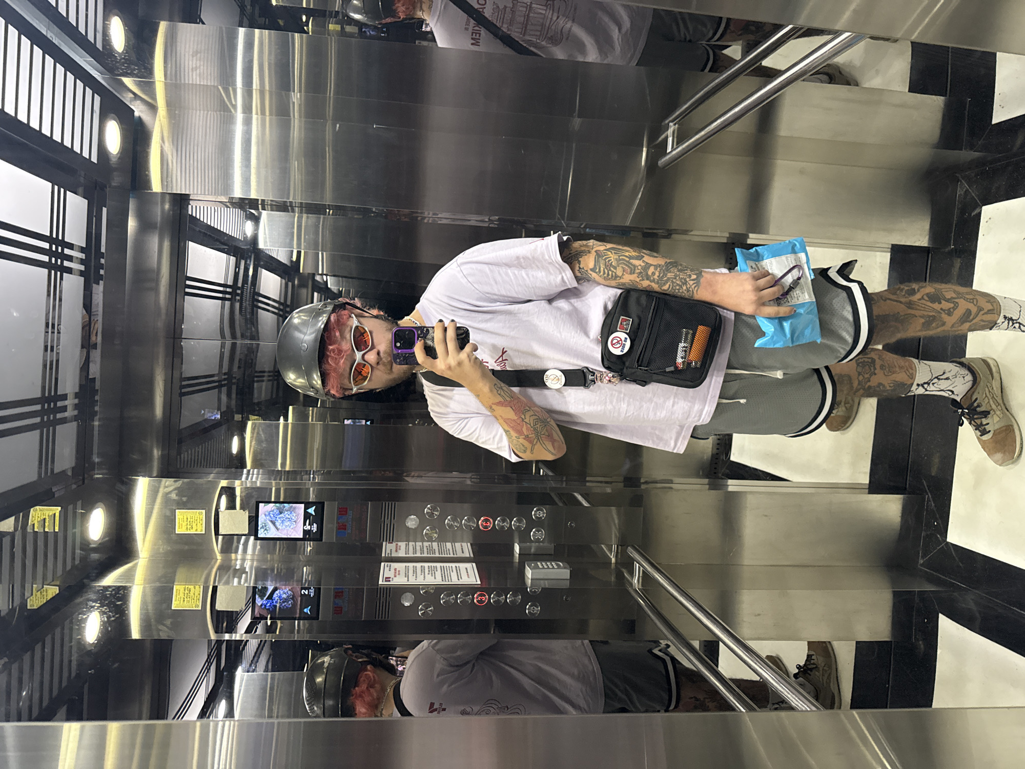 A person in a silver helmet, pink hair, sunglasses, a white t-shirt, gray shorts, and tattooed arms is taking a mirror selfie in an elevator in Đà Nẵng, Vietnam. The elevator has stainless steel walls, a black-and-white tiled floor, and multiple buttons and displays. Another person is reflected in the mirror. The person in the foreground is holding a phone in one hand and a small blue package in the other.