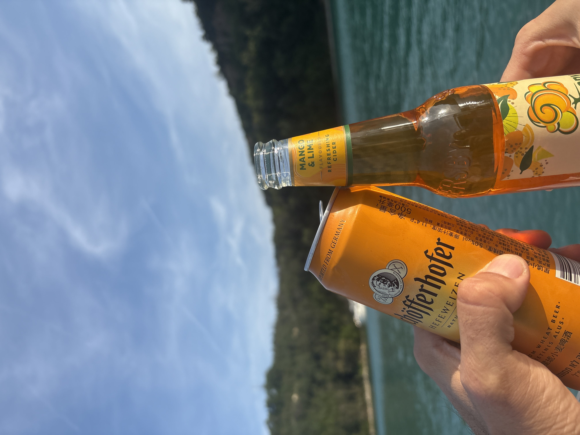 Two human hands are shown holding alcoholic beverages, clinking them together in a toast against an outdoor backdrop. The hand on the left, appearing masculine, grips an orange aluminum can of Schöfferhofer Hefeweizen, a German wheat beer, with text visible in multiple languages including English ("IMPORTED FROM GERMANY"), Chinese, and potentially Hebrew. The can features a circular logo with a bust of a man. The hand on the right, appearing feminine, holds a clear glass bottle containing an orange liquid. Its label reads "MANGO & LIME FLAVOURED REFRESHING CIDER" with fruit illustrations and the partially visible brand "FRUITBY." The bottle is open.
The background consists of a large body of calm, turquoise-blue water in the mid-ground, with dark green, densely vegetated hills rising in the distant background. The sky above is clear and bright blue with scattered, faint wispy white clouds. The scene is illuminated by natural sunlight, suggesting daytime. The location is an unknown body of water flanked by hills, specific city details are not discernible.