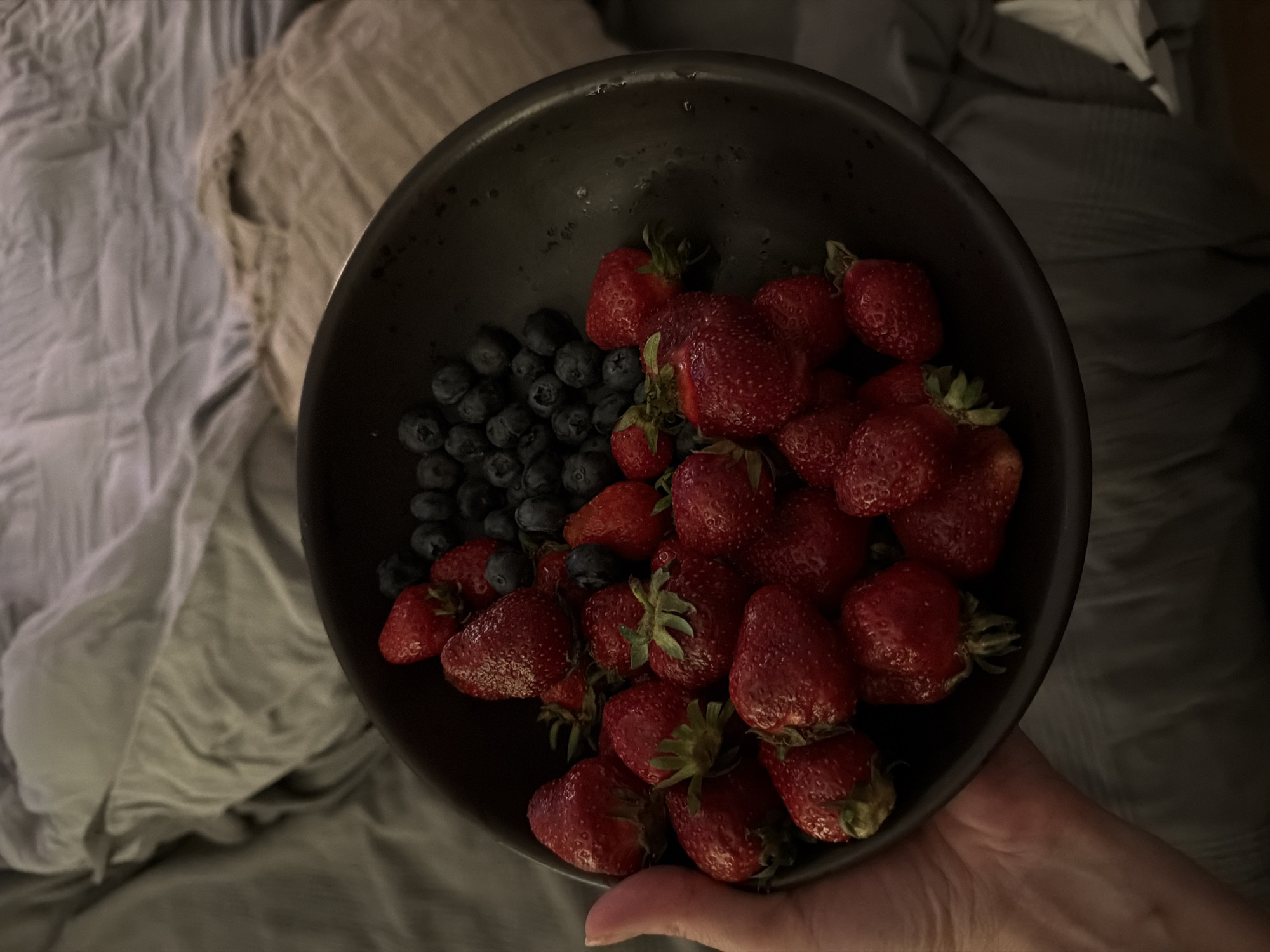 The image presents a close-up, top-down view of a dark-colored bowl filled with fresh berries, held by a person's hand. The bowl contains a large quantity of bright red strawberries, some with their green caps still attached, and a smaller section of dark blue blueberries. Many of the berries appear to be wet, suggesting they have been recently washed.
A hand, with light skin tone, supports the bowl from the lower right side of the frame, with only the thumb and a portion of the palm visible. The background consists of rumpled fabrics, likely bedding or blankets, in shades of grey, beige, and off-white, indicating an indoor setting such as a bedroom.
The lighting is dim and soft, suggesting it might be evening, night, or a dimly lit room. There are no discernible location cues within the image itself to confirm a specific geographic location. No specific activity beyond holding the bowl of fruit is depicted. No text is visible in the media.