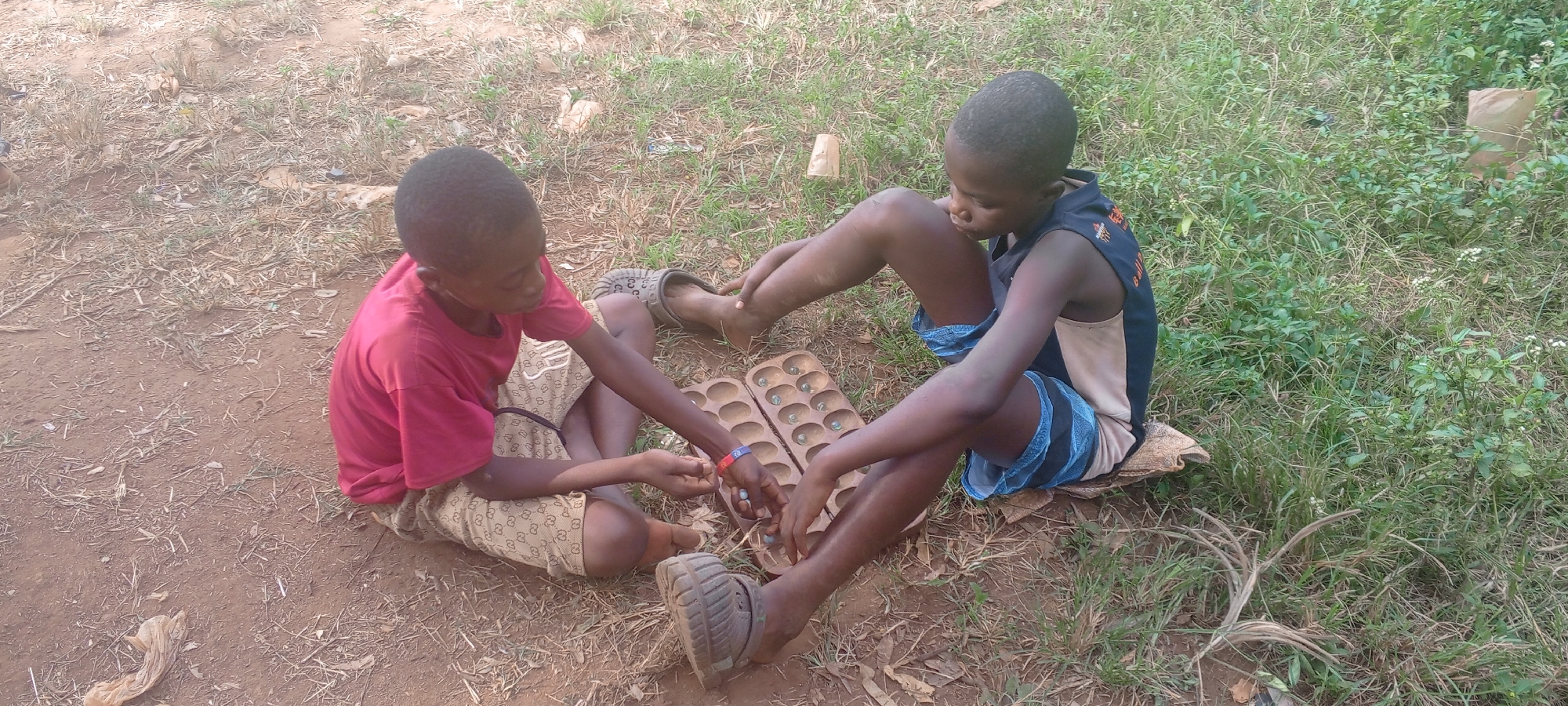 Two young boys are sitting on the ground in an outdoor setting, possibly in Ngalula, Democratic Republic of the Congo. The boy on the left is wearing a red t-shirt and patterned shorts. The boy on the right is wearing a sleeveless shirt and blue shorts. They are playing a game of Mancala with a wooden board placed between them. The ground is a mix of dirt, grass, and some scattered vegetation. There are also a few discarded items visible in the surrounding area.